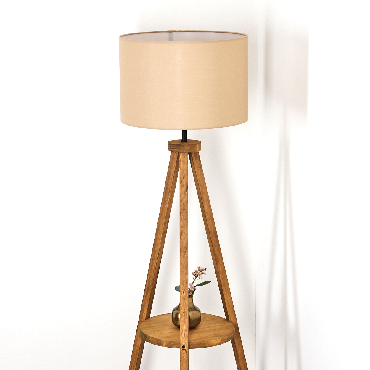 Eine Stehlampe aus Holz mit beigem Lampenschirm. Eine kleine Vase mit Blumen steht auf einem runden Regal. Weißer Wandhintergrund.