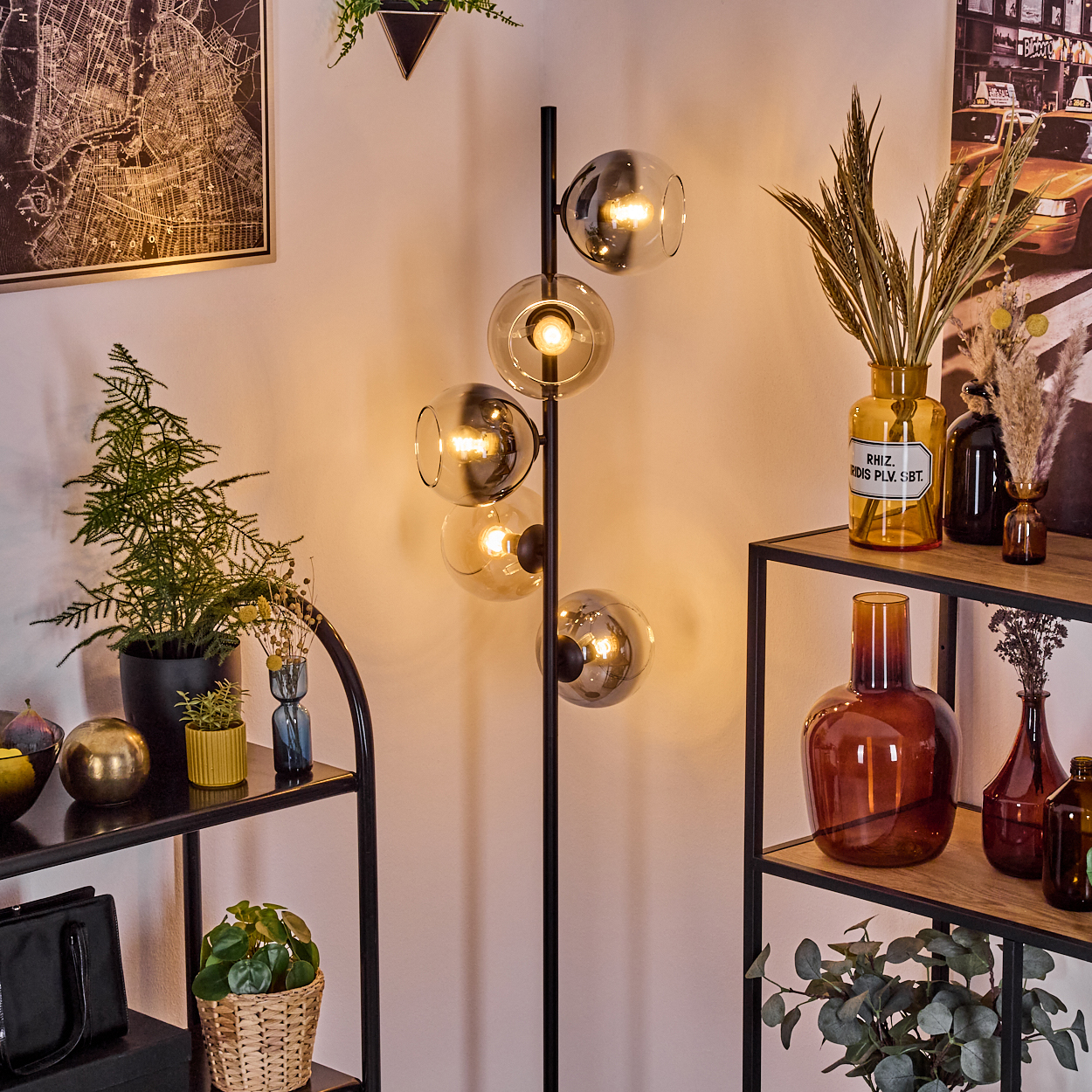 Eine Zimmerecke mit Stehlampe. Regale mit Vasen und Pflanzen stehen rechts und links. Eine Karte hängt an der linken Wand.