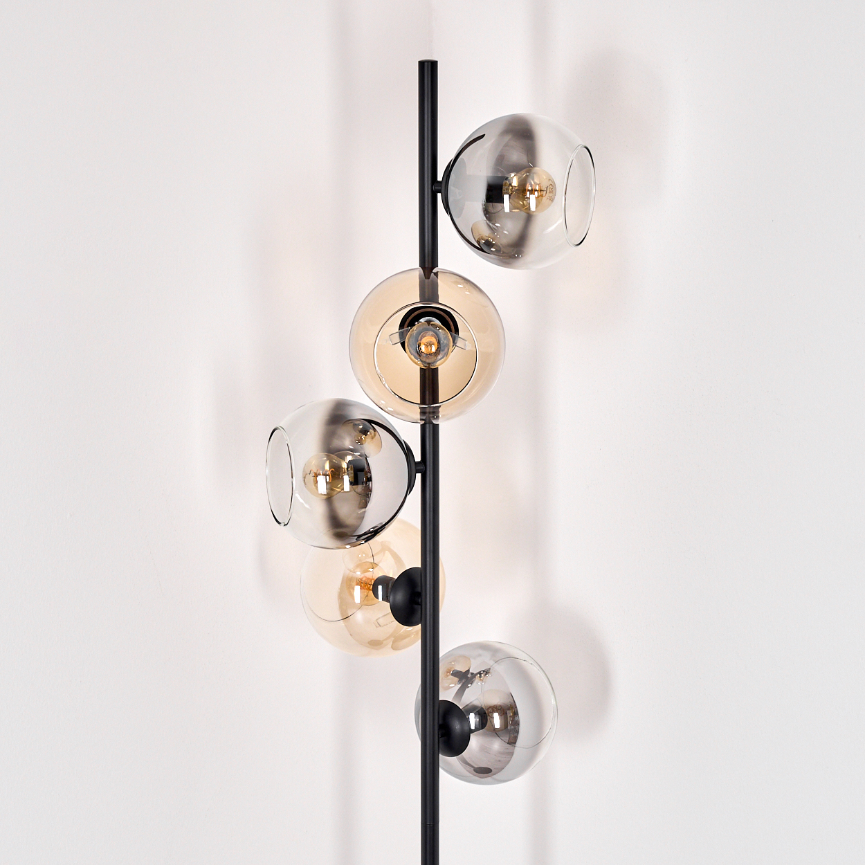 Eine moderne Stehlampe mit fünf Glaskugelschirmen, abwechselnd klar und bernsteinfarben, an einem schwarzen Metallpfahl, vor einer weißen Wand.
