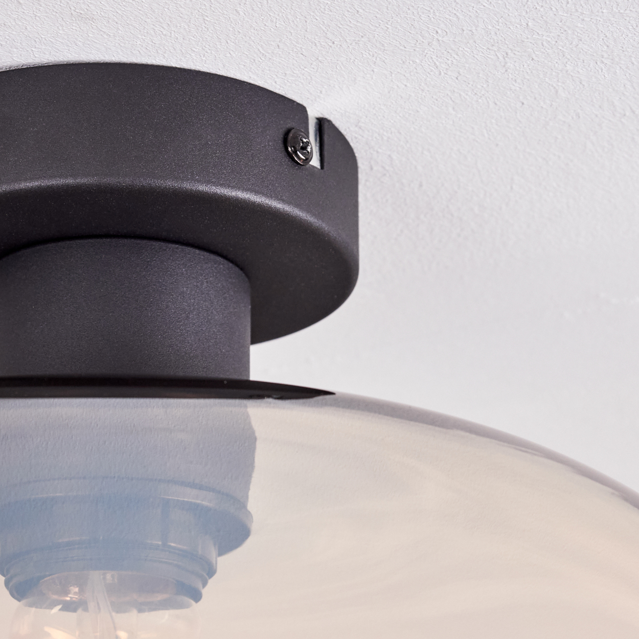 Nahaufnahme einer schwarzen Deckenlampe. Die Lampe hat einen dunklen Sockel und eine transparente Glaskuppel. Der Hintergrund ist eine weiße, strukturierte Decke.