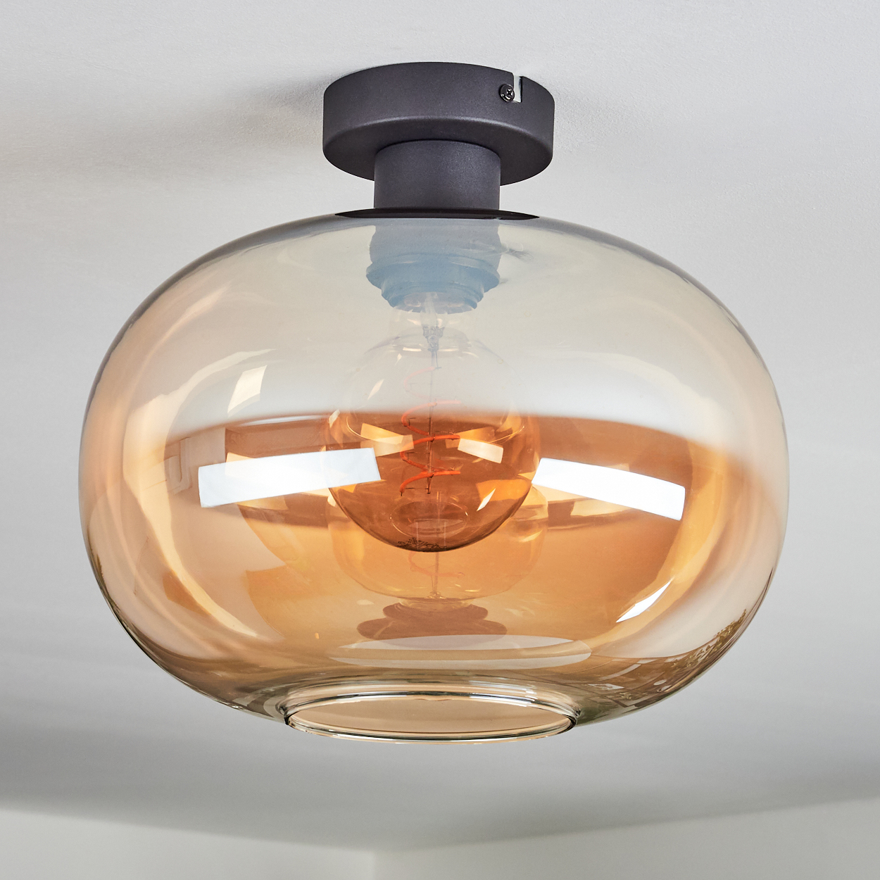 Eine moderne Deckenleuchte mit rundem, bernsteinfarbenem Glasschirm. Im Inneren befindet sich eine sichtbare Glühbirne. Die Leuchte ist an einer weißen Decke befestigt.
