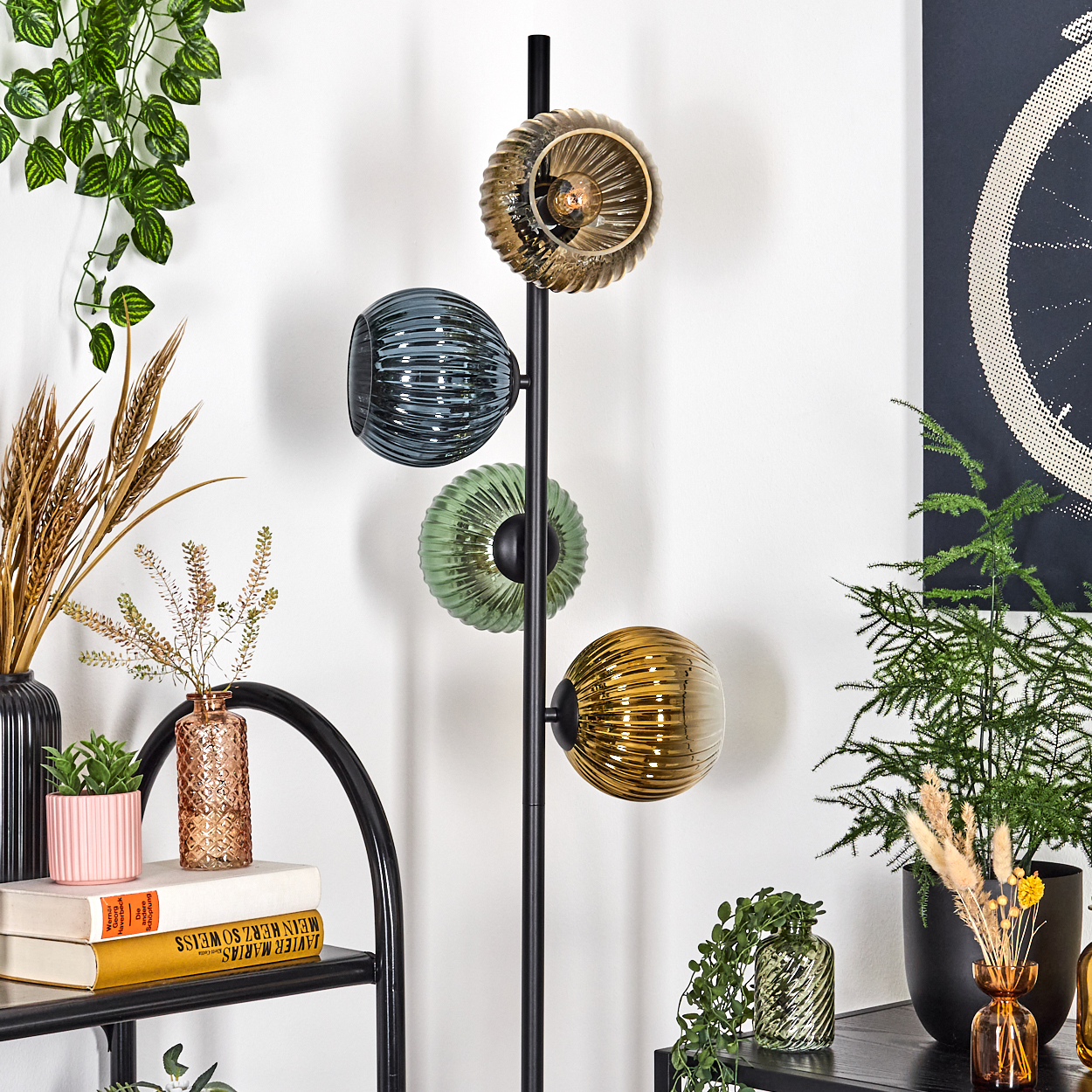 Eine moderne Lampe mit vier farbigen Glaskugeln. Pflanzen und Dekorationsartikel umgeben sie auf Regalen, mit einem Fahrradrad-Kunstwerk im Hintergrund.