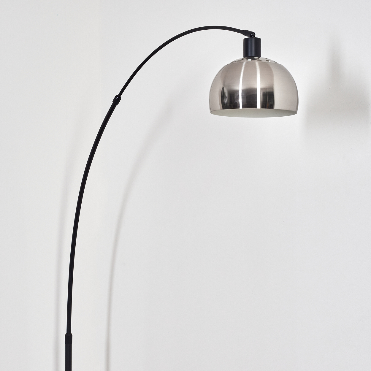 Eine moderne Bogenlampe mit silbernem Lampenschirm und schwarzem Arm steht vor einer weißen Wand. Der Arm ist geschwungen.