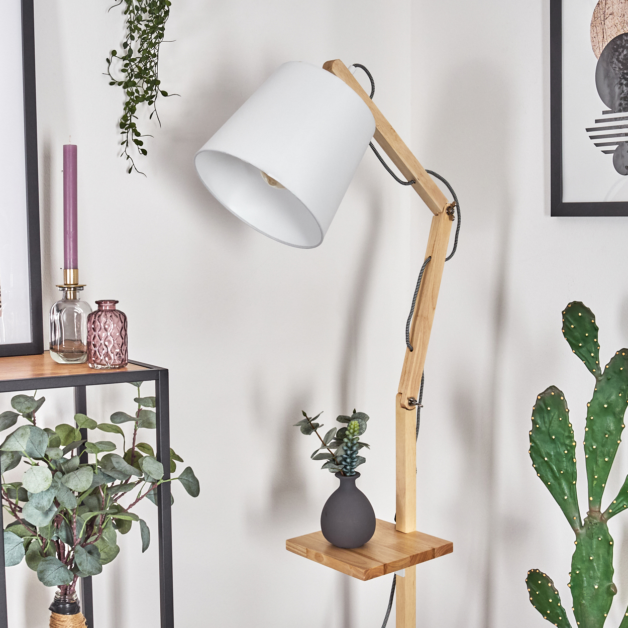 Eine moderne Stehlampe aus Holz mit weißem Lampenschirm. Ein kleines Regal hält eine dunkle Vase mit Pflanzen. In der Nähe steht ein hoher Kaktus.