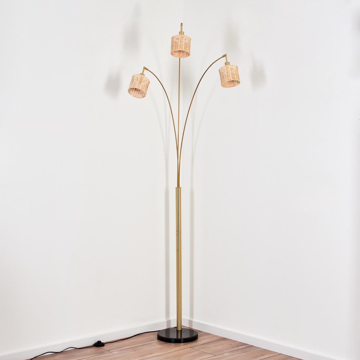 Eine goldene Stehlampe mit drei Rattanschirmen steht in einer Ecke. Der Sockel ist schwarz, die Wände sind weiß und der Boden ist Holz.