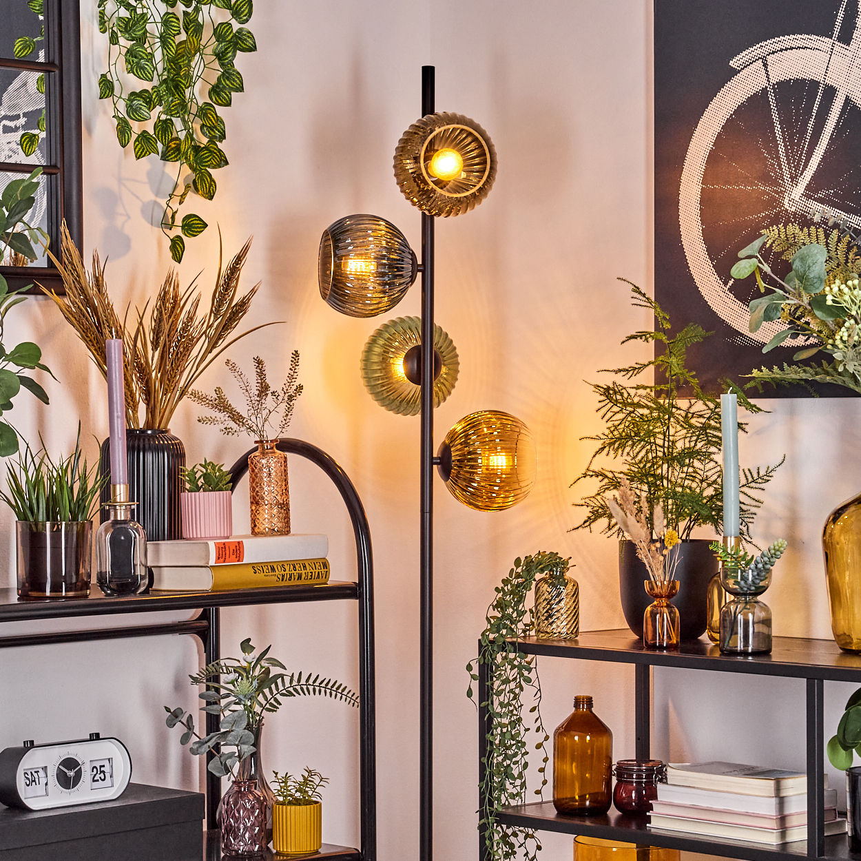 Ein Raum mit einer Stehlampe und Regalen mit Pflanzen, Vasen, Büchern und einer Uhr. Im Hintergrund hängt ein Fahrradrad-Kunstwerk.