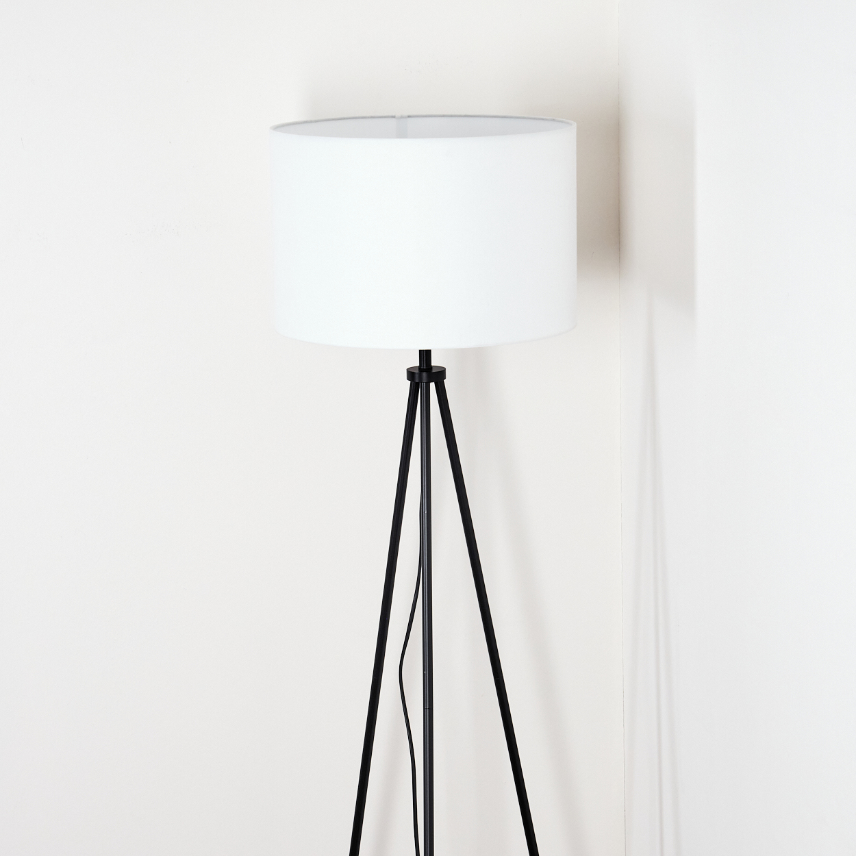 Ein weißer Lampenschirm sitzt auf einer schwarzen Stativlampe. Die Lampe steht in einer Ecke mit weißen Wänden.