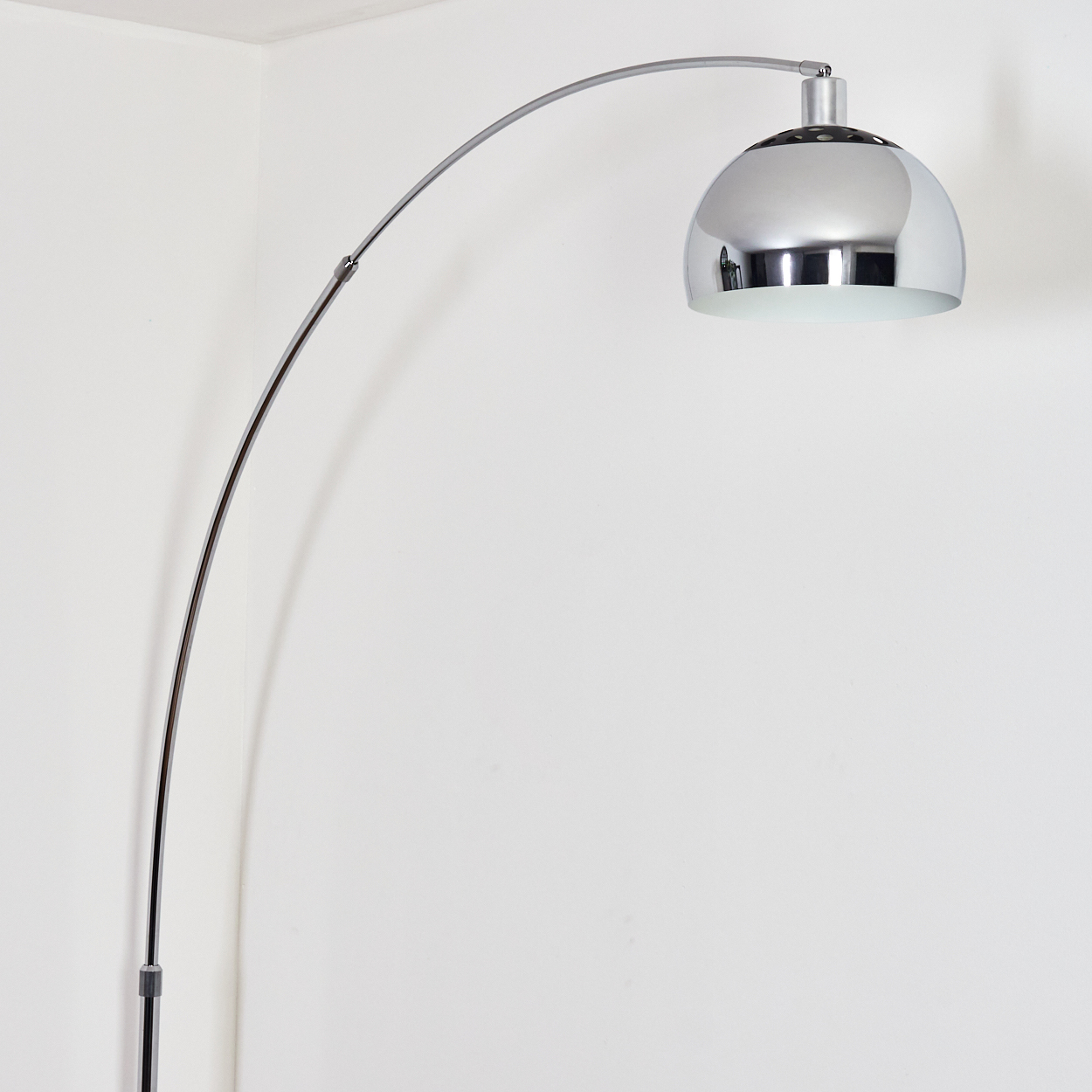 Eine verchromte Bogen-Stehlampe. Sie hat einen gebogenen Arm, einen gewölbten Lampenschirm und ein weißes Innere, vor einer weißen Wand.