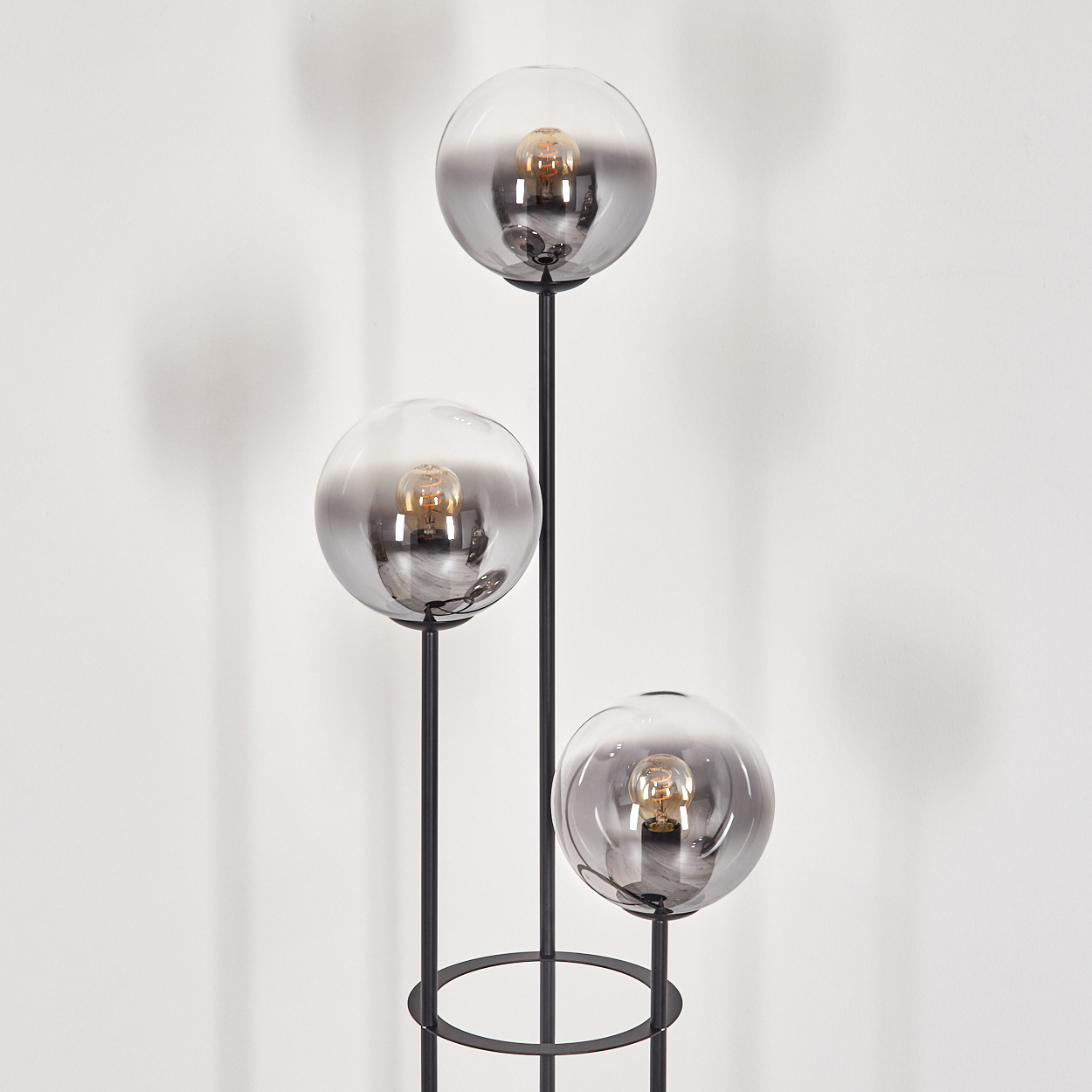 Eine moderne Stehlampe mit drei kugelförmigen Schirmen. Jede hat eine Glühbirne. Die Lampe ist schwarz mit rundem Fuß und weißer Wand im Hintergrund.