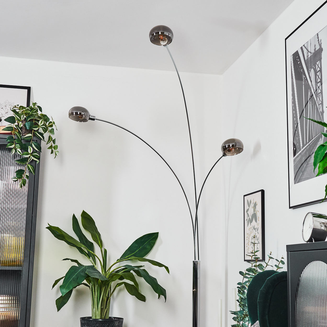 Eine moderne Stehlampe mit drei Kugelleuchten. Pflanzen und gerahmte Kunstwerke im Hintergrund, eine Uhr auf einem schwarzen Schrank.
