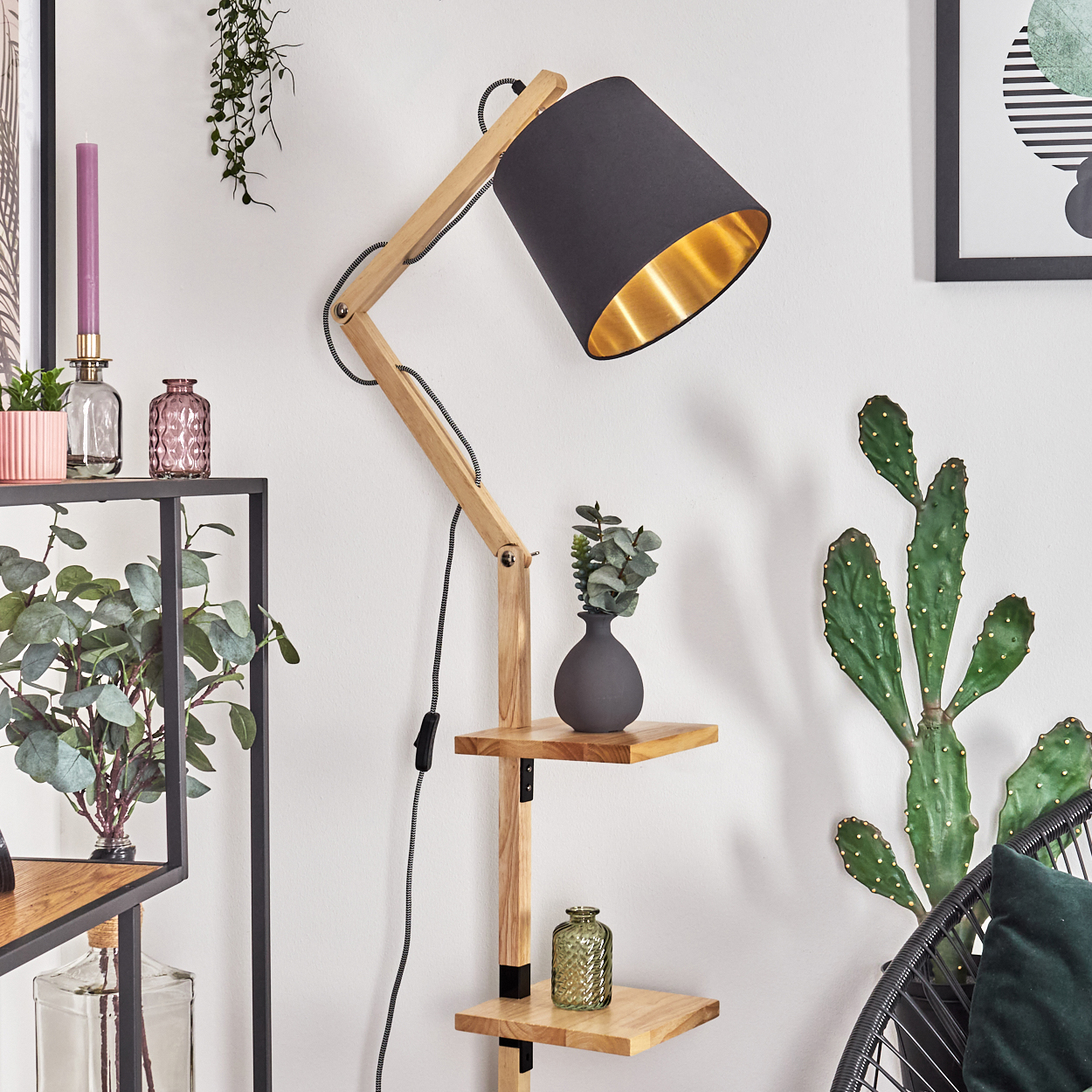 Eine Holzlampe mit schwarzem Schirm und goldenem Innenraum. Ein Regal mit Vasen und Pflanzen befindet sich in der Nähe. Ein Kaktus und ein gerahmtes Kunstwerk sind im Hintergrund.