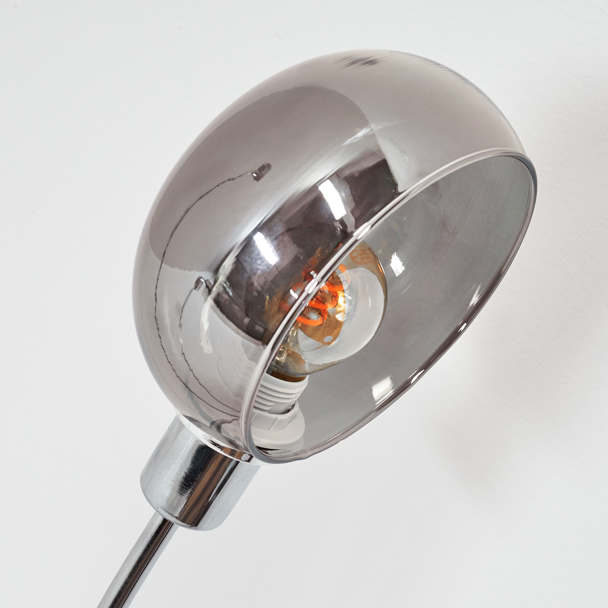 Eine Chromlampe mit einer Glühbirne in der Mitte. Die Lampe hat einen gebogenen Schirm und ein glänzendes, metallisches Finish vor weißem Hintergrund.