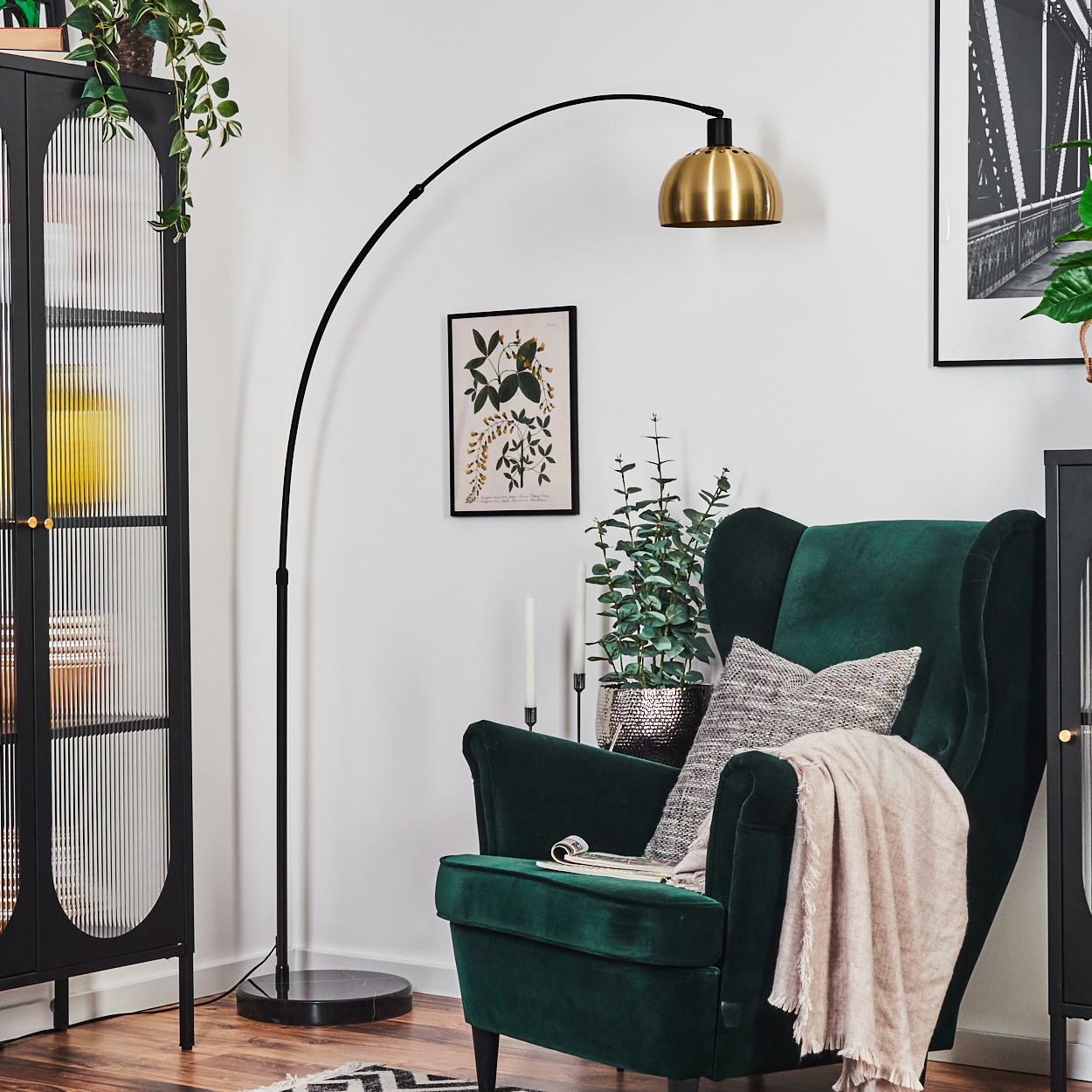 Ein grüner Sessel mit Decke und Kissen, in der Nähe einer Stehlampe und eines schwarzen Schranks. Eine Pflanze und ein Bild sind ebenfalls vorhanden.