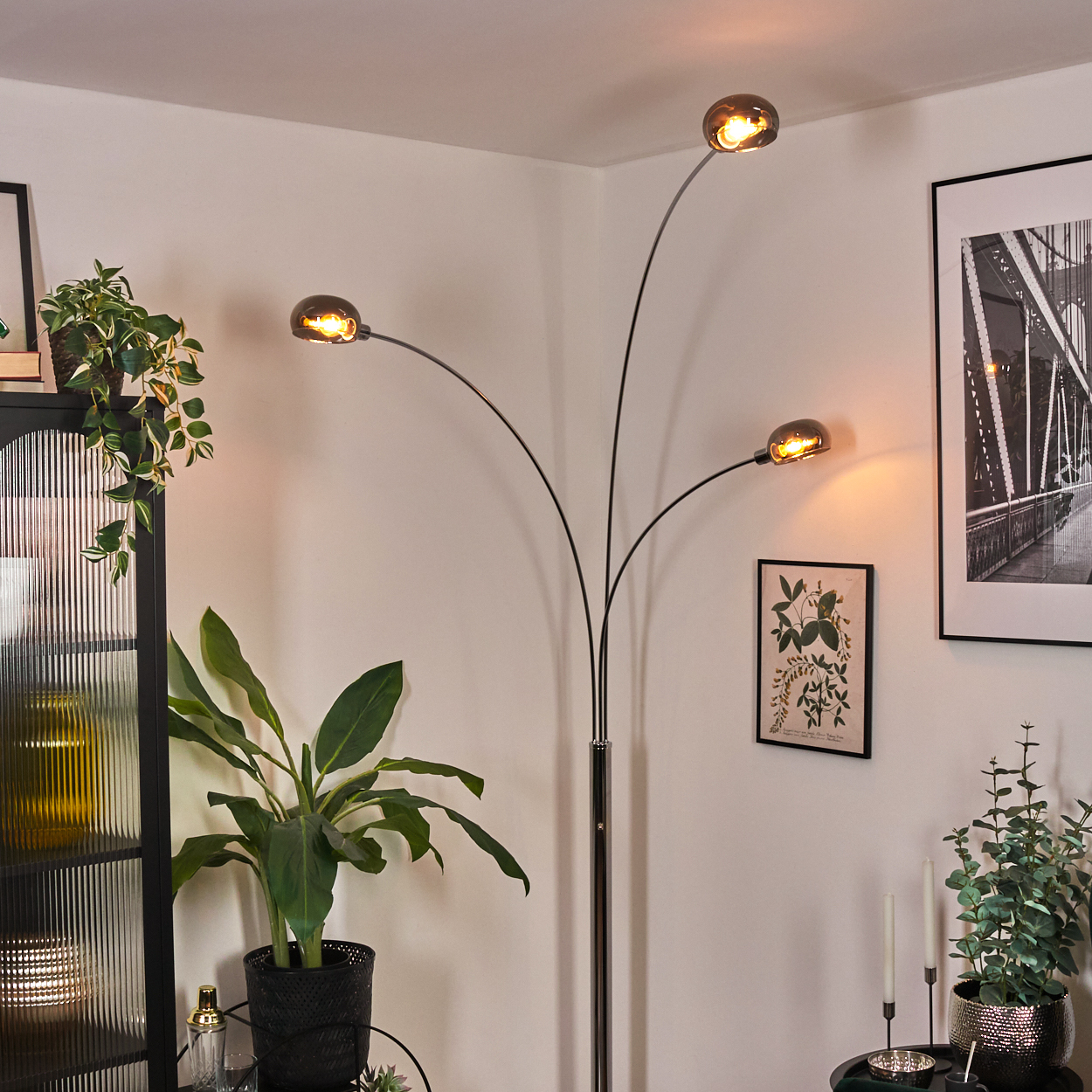 Eine moderne Stehlampe mit drei Lichtern. Pflanzen und ein gerahmtes Brückenfoto sind sichtbar. Die Szene ist drinnen mit weißen Wänden.