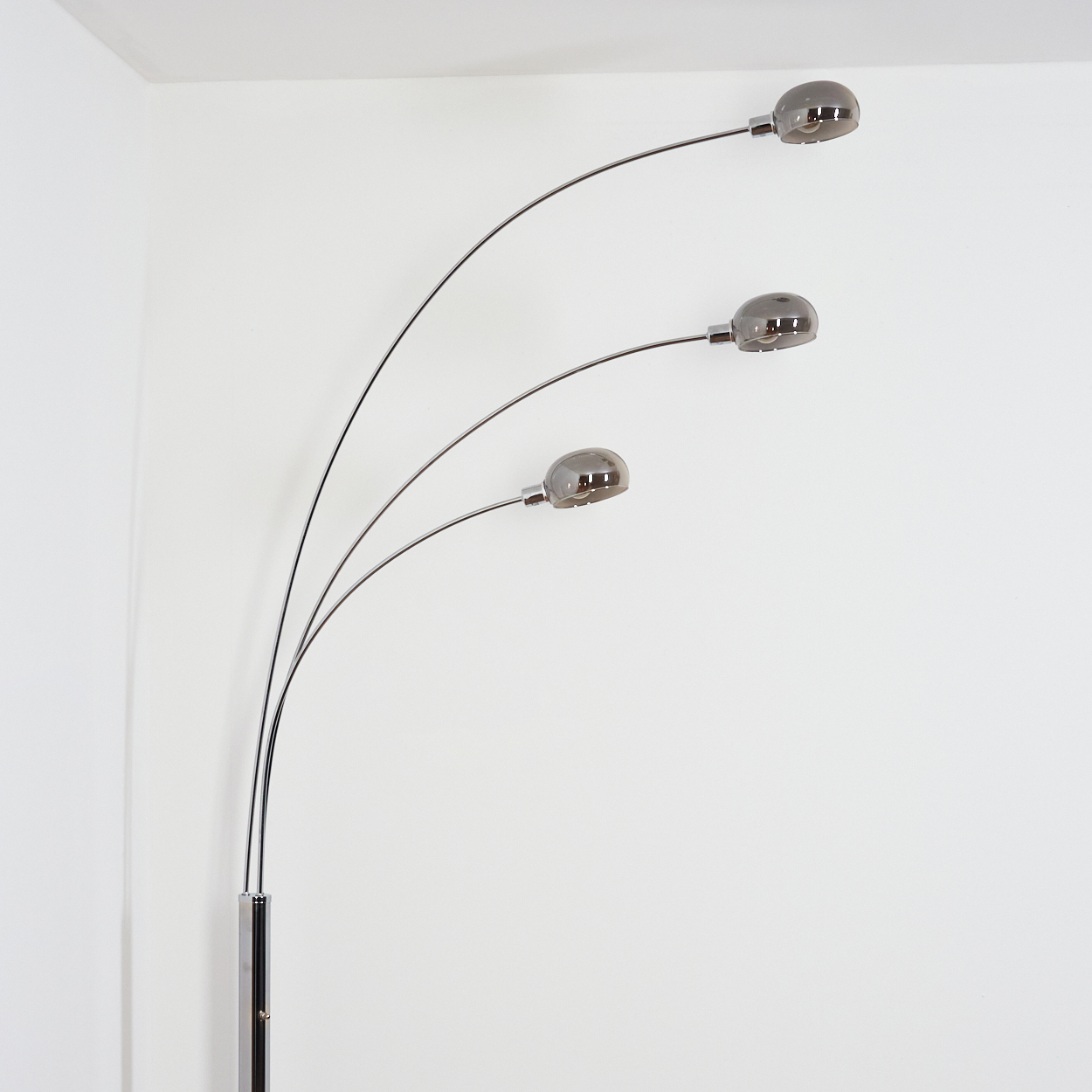 Eine moderne, verchromte Stehlampe mit drei gebogenen Armen, die jeweils eine graue, kuppelförmige Leuchte tragen. Sie steht vor einer weißen Wand.