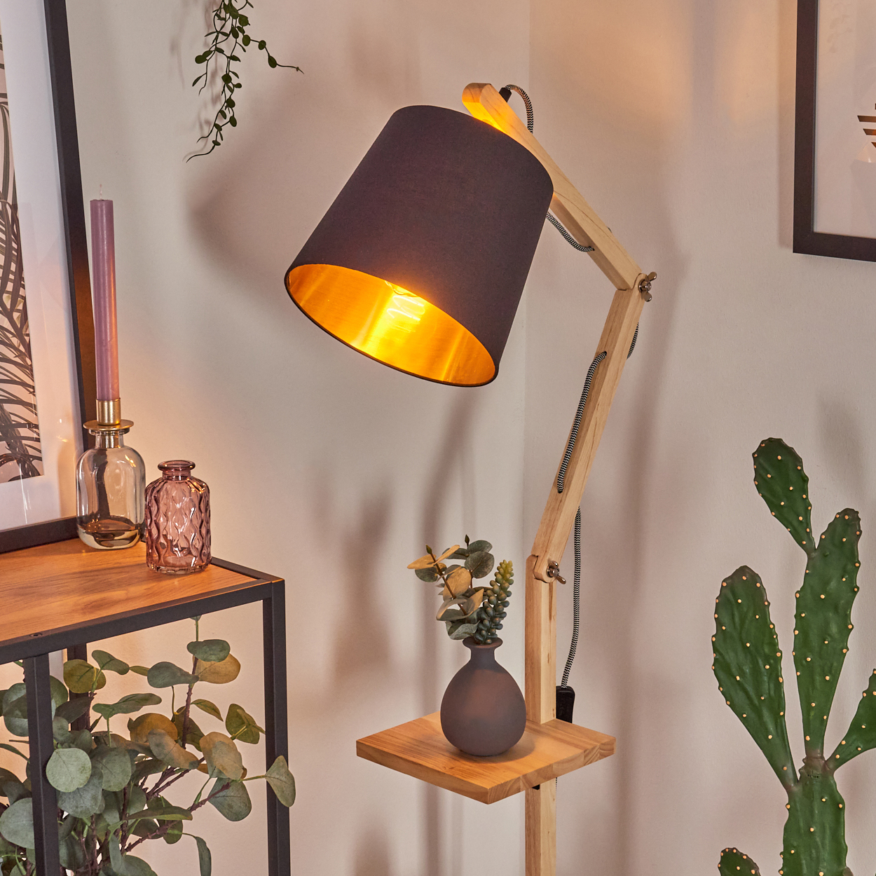 Moderne Stehlampe aus Holz mit schwarzem Lampenschirm, neben Kaktus, Regal mit Pflanzen und Tisch mit Kerze und Vasen.