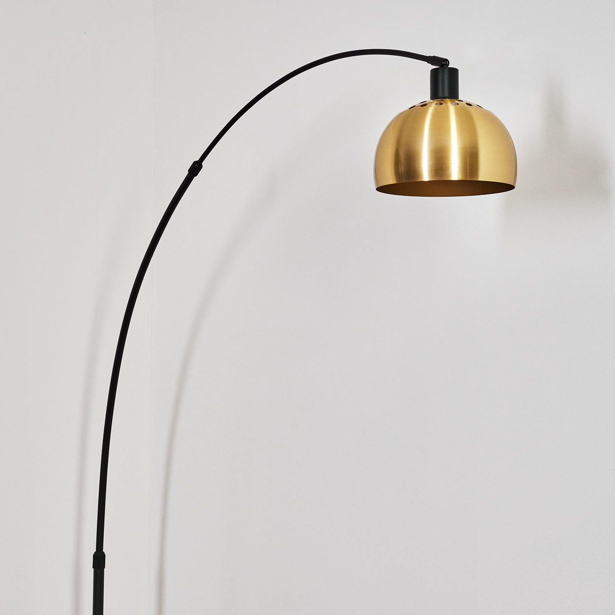 Eine moderne Bogenstehlampe. Sie hat einen schwarzen, gebogenen Arm und einen goldenen, glockenförmigen Lampenschirm vor einer weißen Wand.