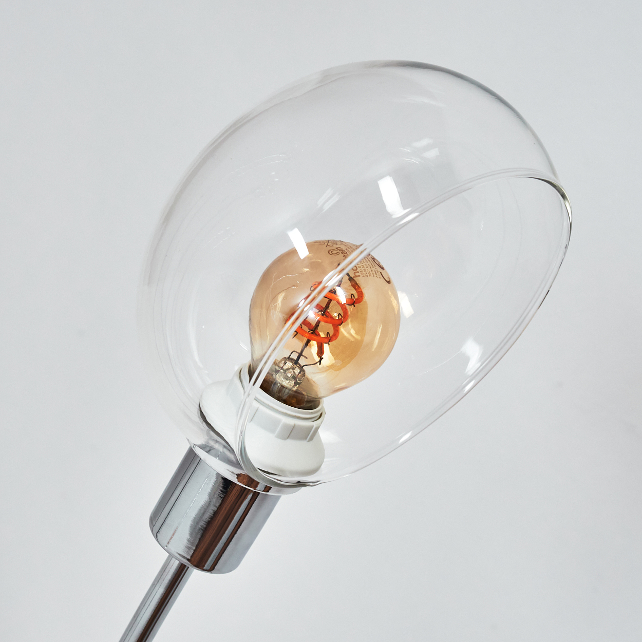 Eine moderne Lampe mit einem durchsichtigen Glasschirm und einer warm getönten Glühbirne. Die Lampe hat einen Chromschaft vor weißem Hintergrund.