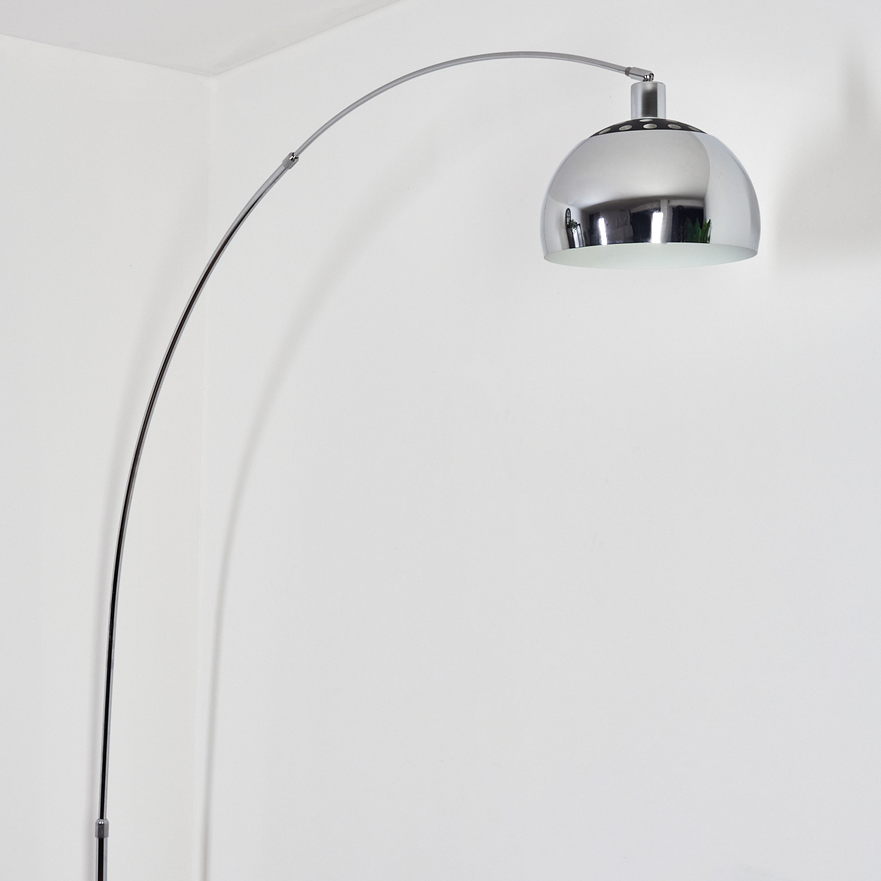 Eine verchromte Bogenlampe mit rundem Schirm. Sie steht vor einer weißen Wand und Ecke. Die Lampe spiegelt die Umgebung wider.