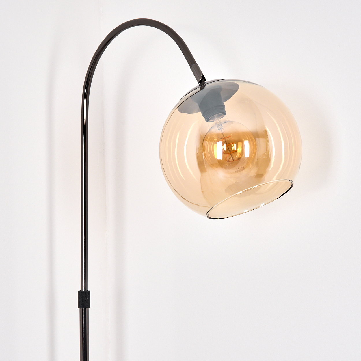 Eine moderne Stehlampe mit dunklem Metallrahmen und einem bernsteinfarbenen Glaskugel-Schirm, mit einer leuchtenden Glühbirne. Der Hintergrund ist weiß.