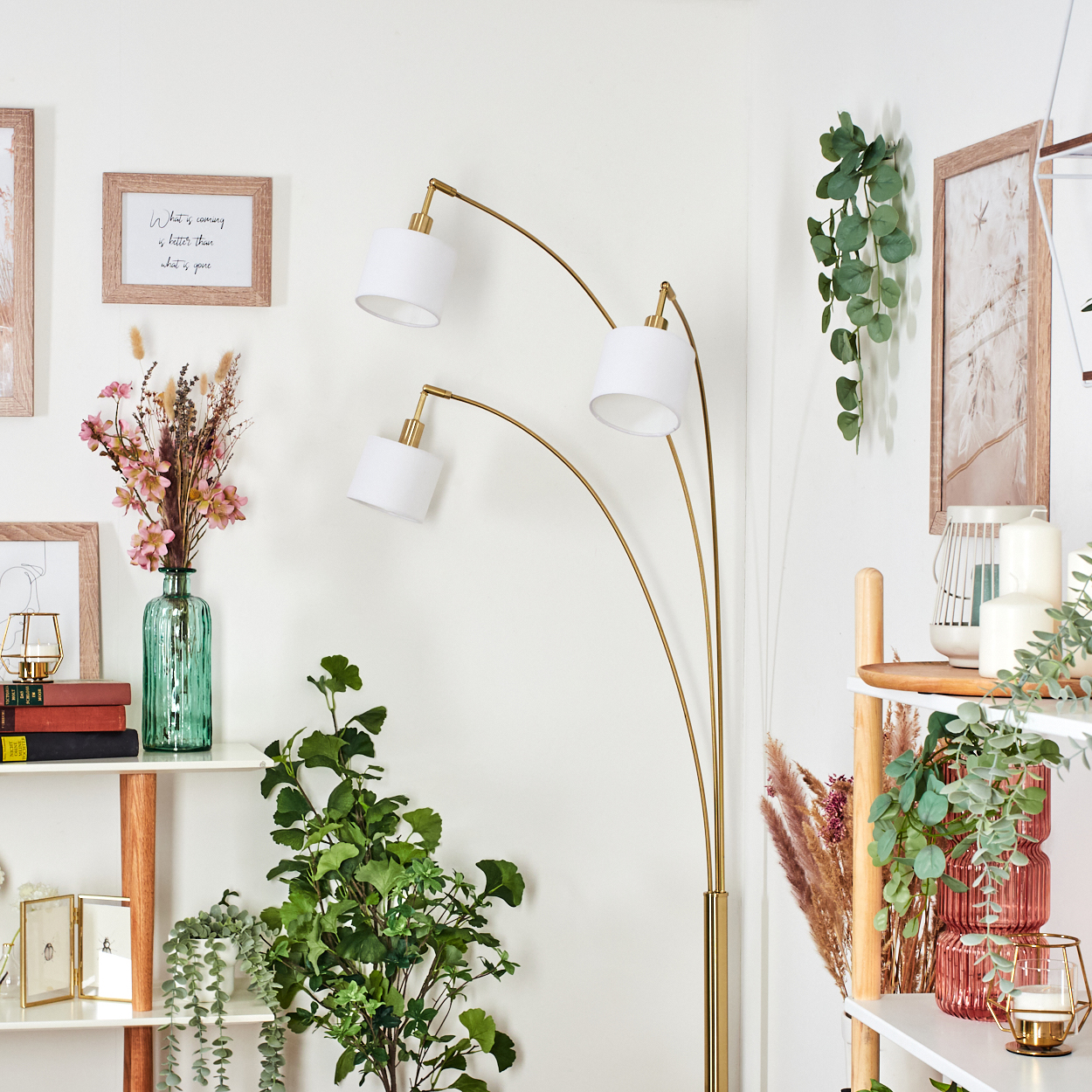 Ein Raum mit einer goldenen Lampe, Regalen mit Pflanzen und Dekoration. Eine grüne Vase mit Blumen und Bilder an der Wand sind zu sehen.