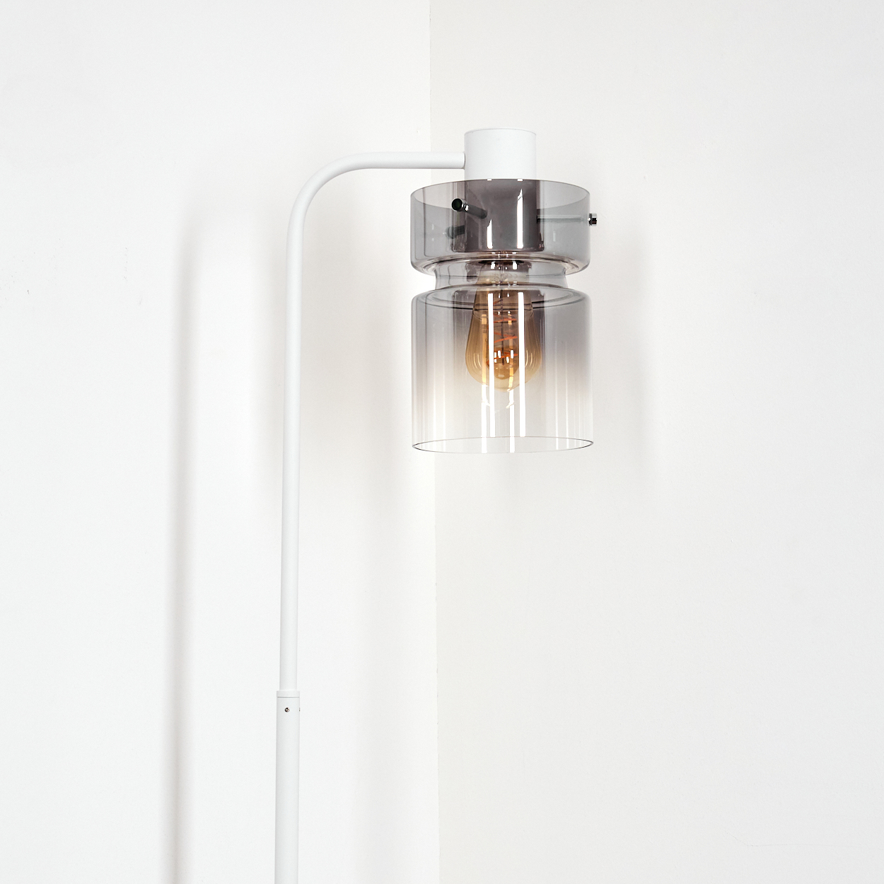 Eine moderne weiße Stehlampe mit Glasschirm und Glühbirne. Sie steht vor einer weißen Wand.