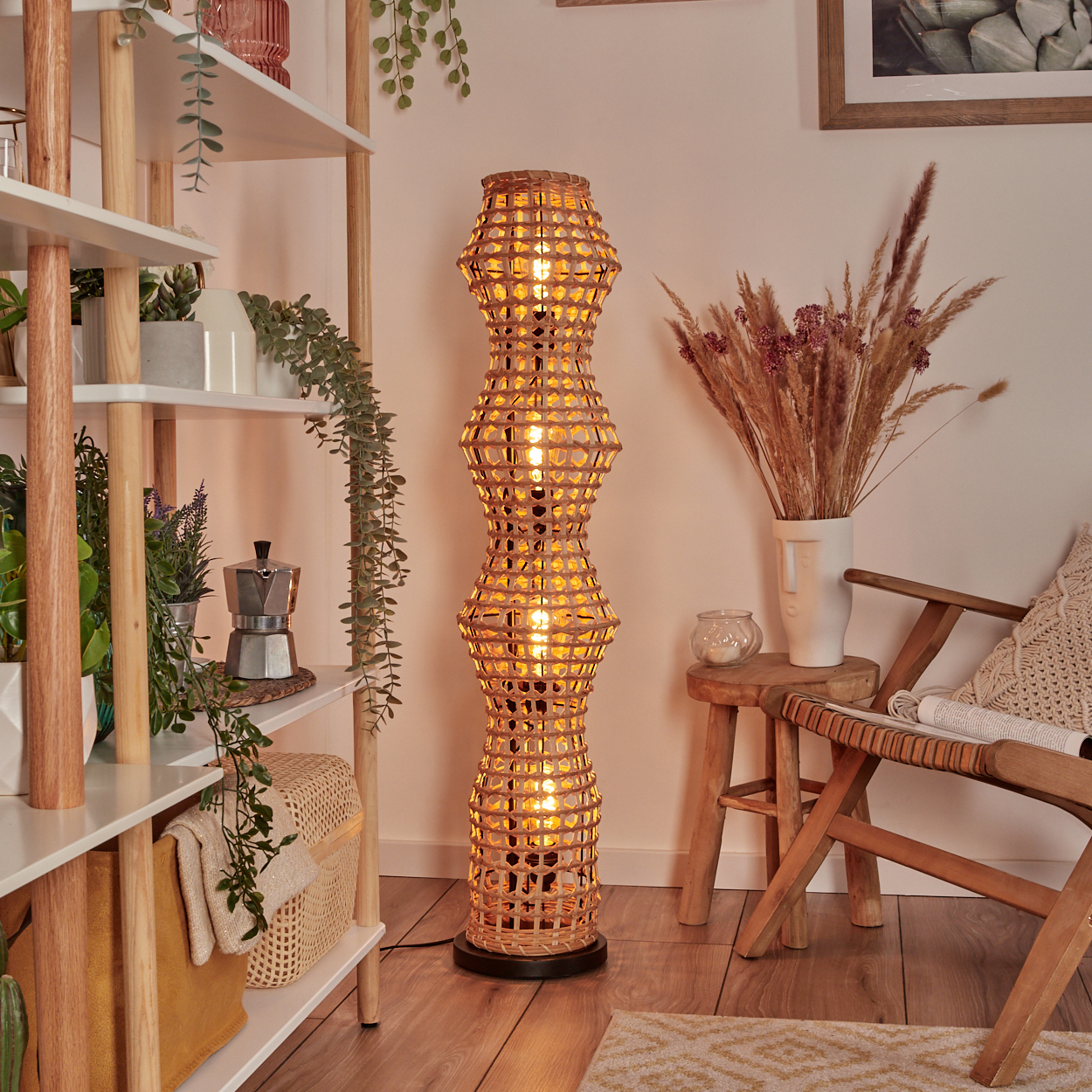 Eine gewebte Stehlampe steht in einem Raum mit Regalen, Pflanzen und einem Holzstuhl. Trockenblumen stehen in einer weißen Vase.