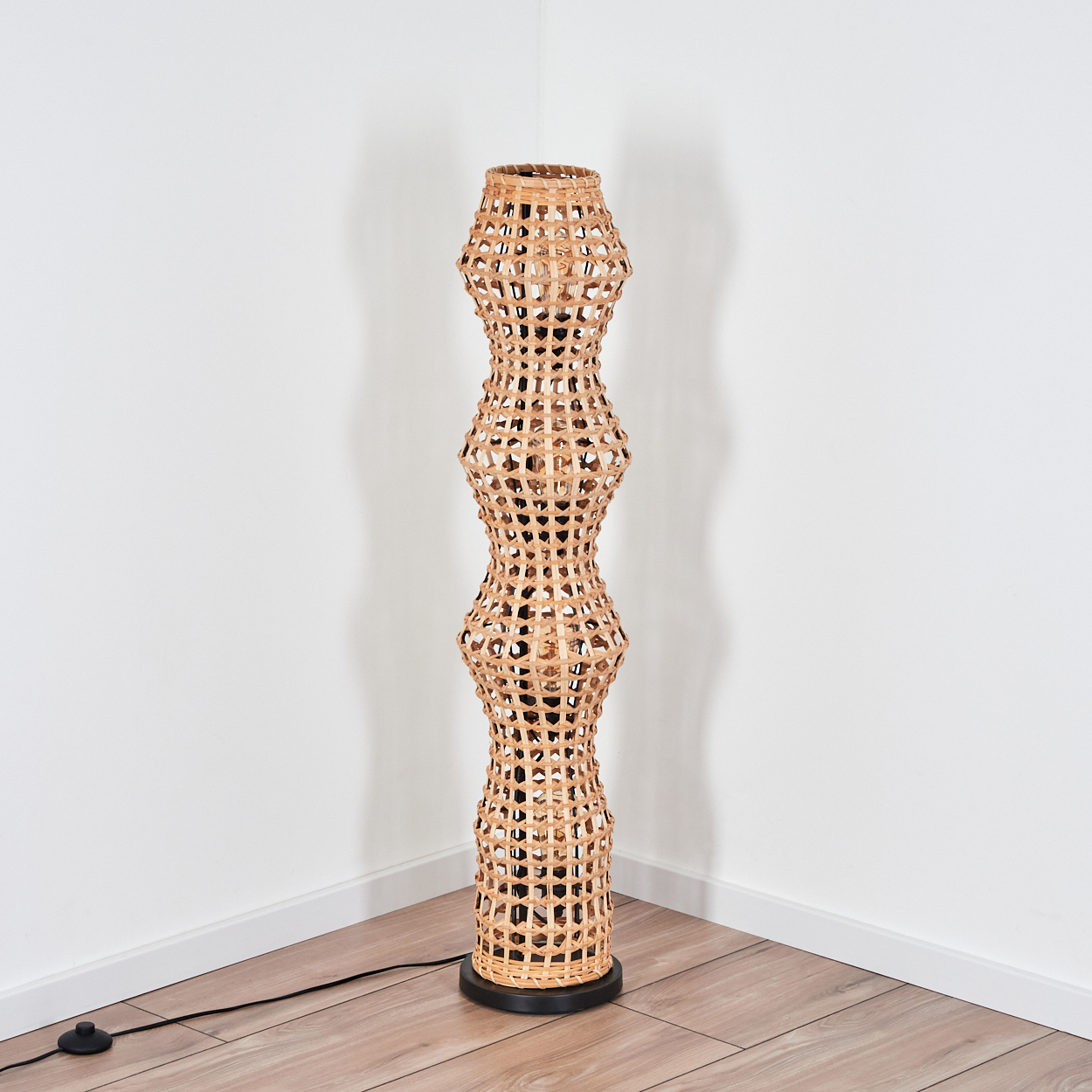Eine hohe, gewebte Stehlampe aus Rattan steht in einer Ecke. Sie ist beige und schwarz, mit einem schwarzen Sockel und einem Netzkabel.