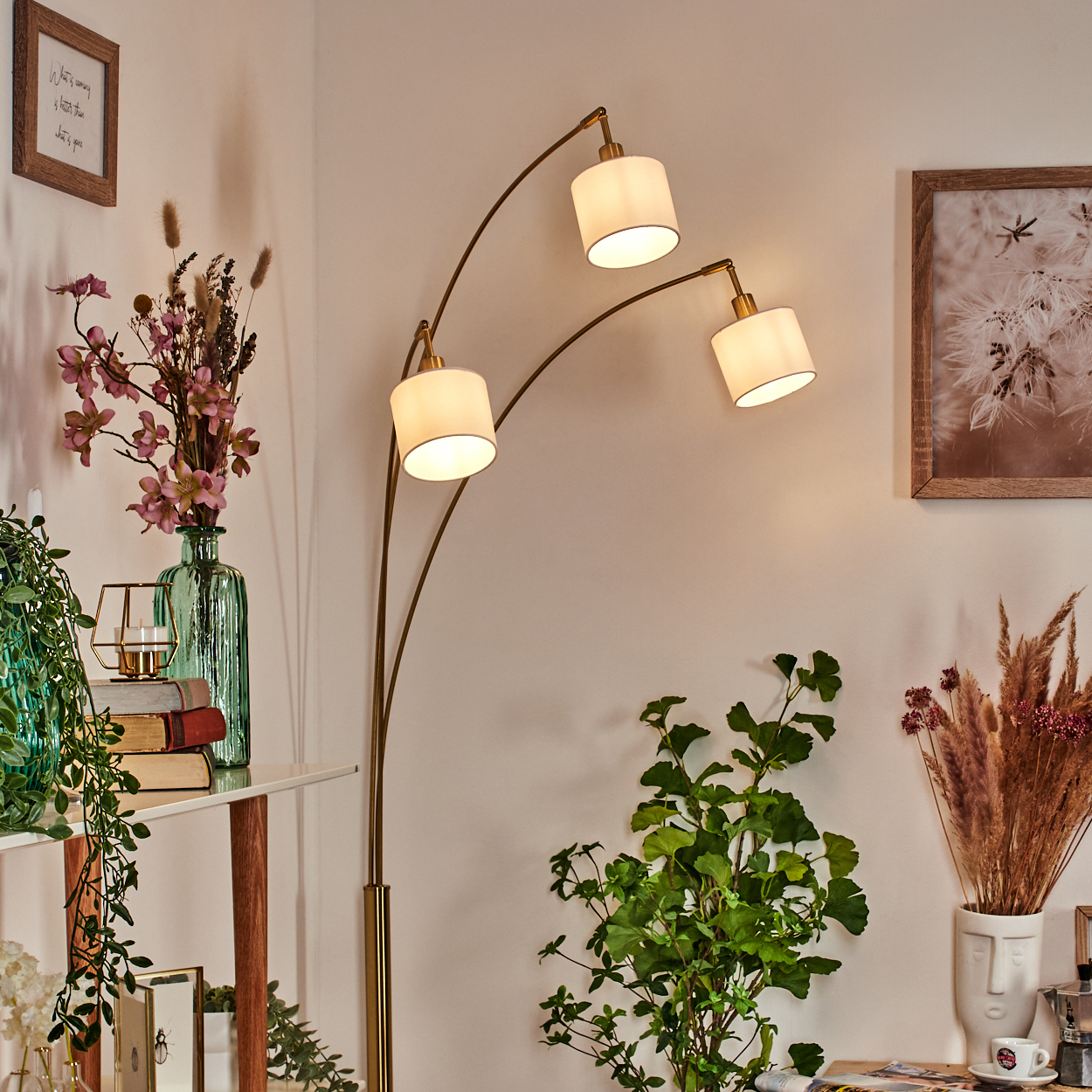 Ein Raum mit einer dreifach-Lampen-Stehlampe, Pflanzen, Vasen, Büchern und gerahmten Bildern an der Wand. Der Raum ist warm beleuchtet.
