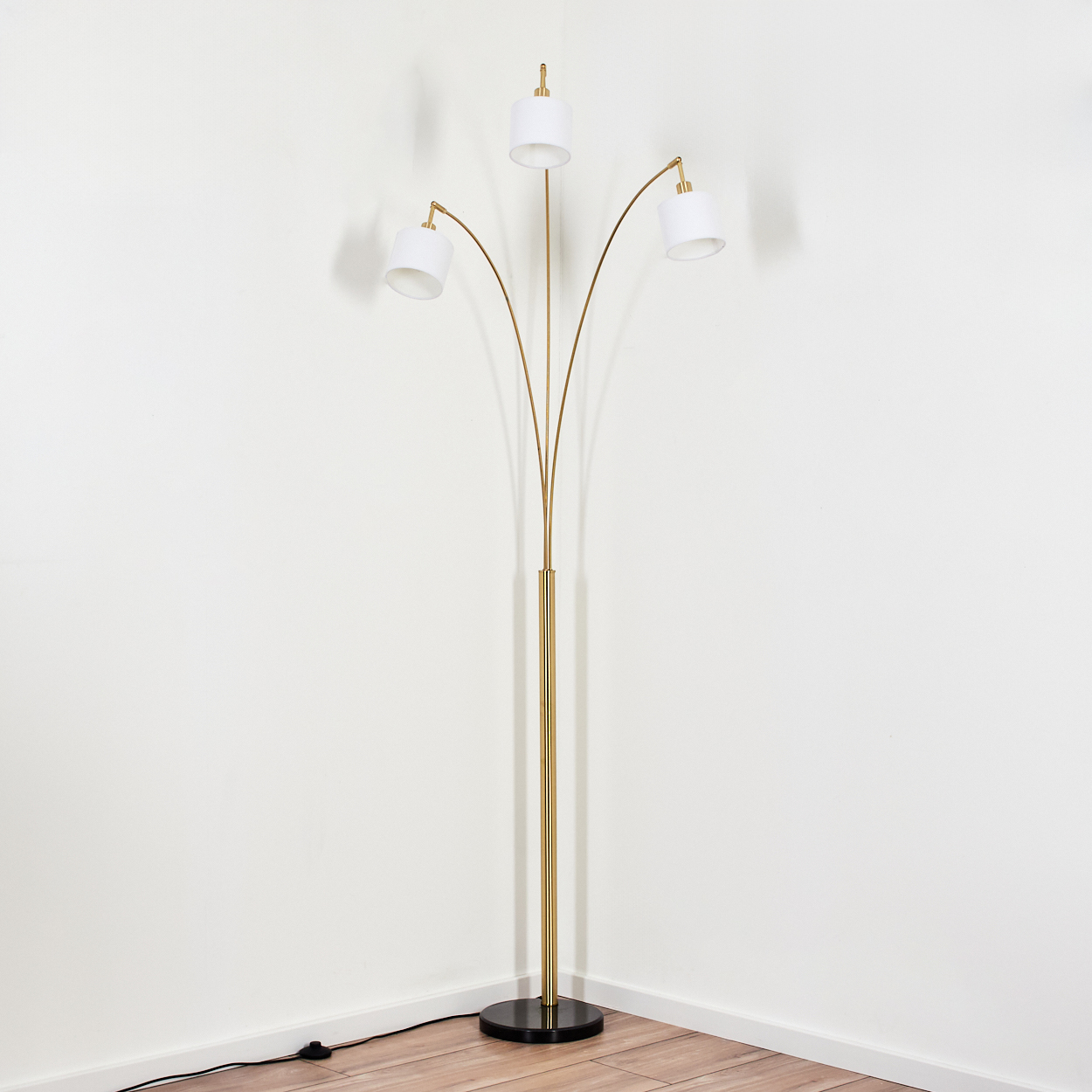 Eine goldene Stehlampe mit drei weißen Schirmen. Sie steht in einer Ecke mit weißen Wänden und Holzboden. Ein schwarzer Sockel ist sichtbar.