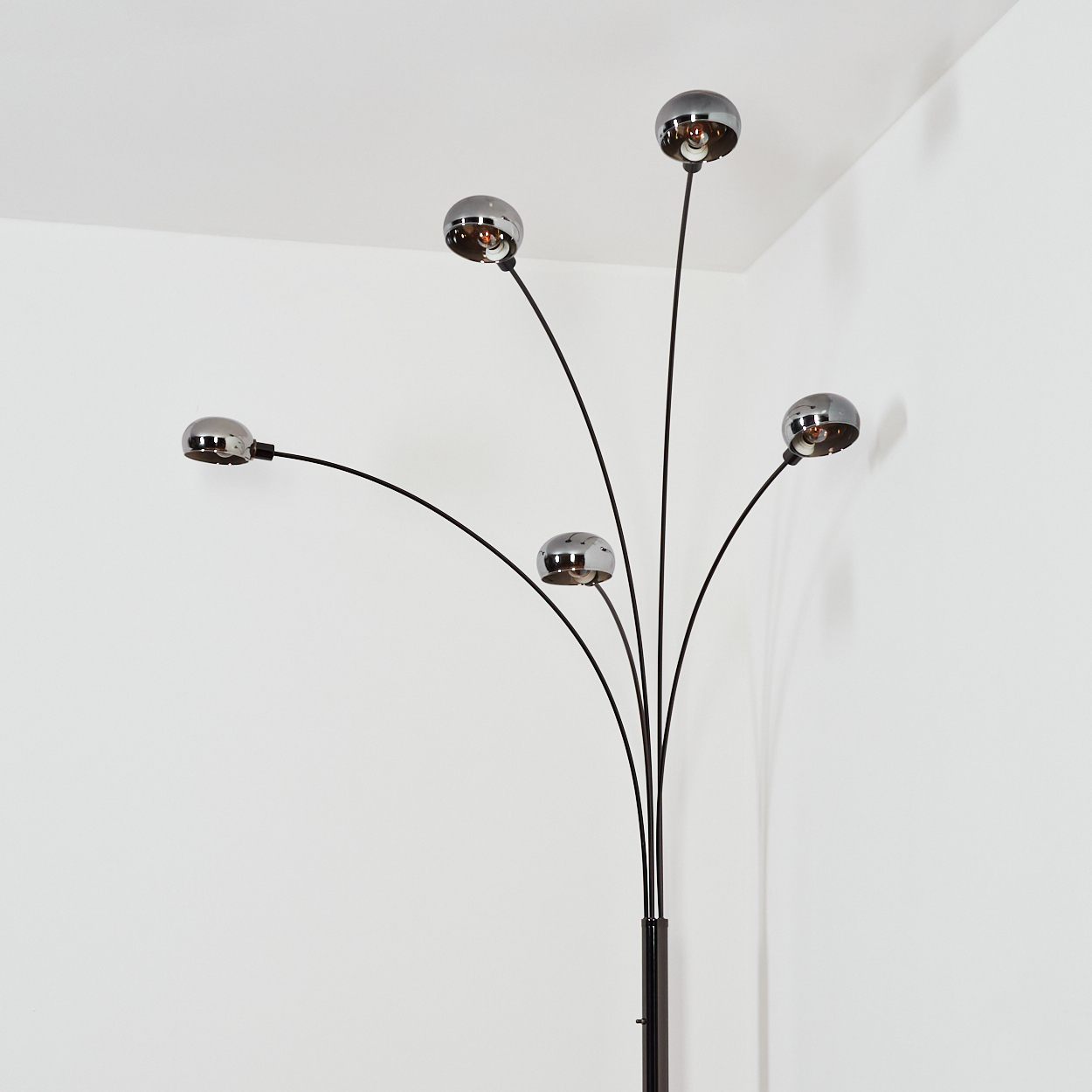 Eine moderne Stehlampe mit fünf Chrom-Kugelleuchten, schwarzen Stielen und einem dunklen Sockel vor einer weißen Wand in einer Ecke.