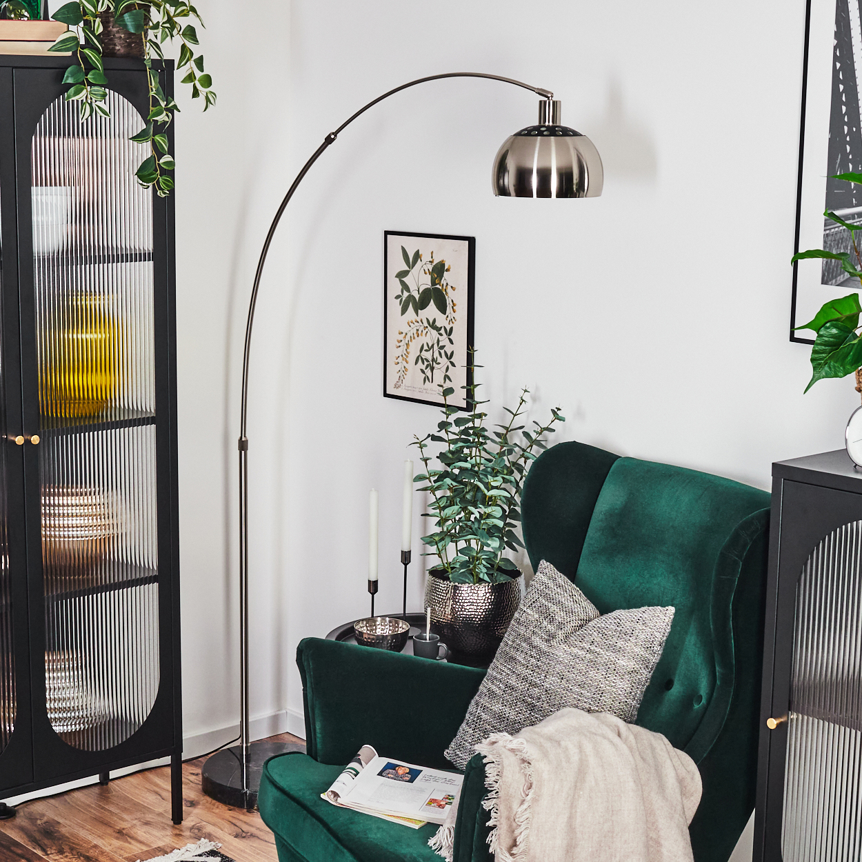 Ein grüner Sessel mit Kissen, eine Stehlampe, ein schwarzer Schrank und Pflanzen. Auf dem Stuhl liegt eine Zeitschrift.