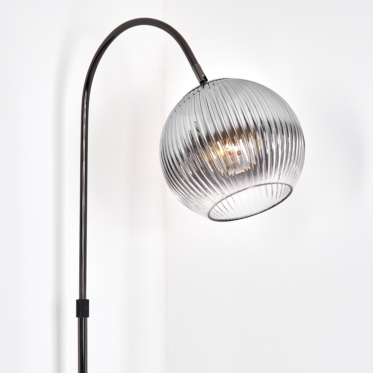 Eine moderne Stehlampe mit einem klaren, geriffelten Glaskugel. Die Lampe hat einen schwarzen Metallrahmen und steht an einer weißen Wand.