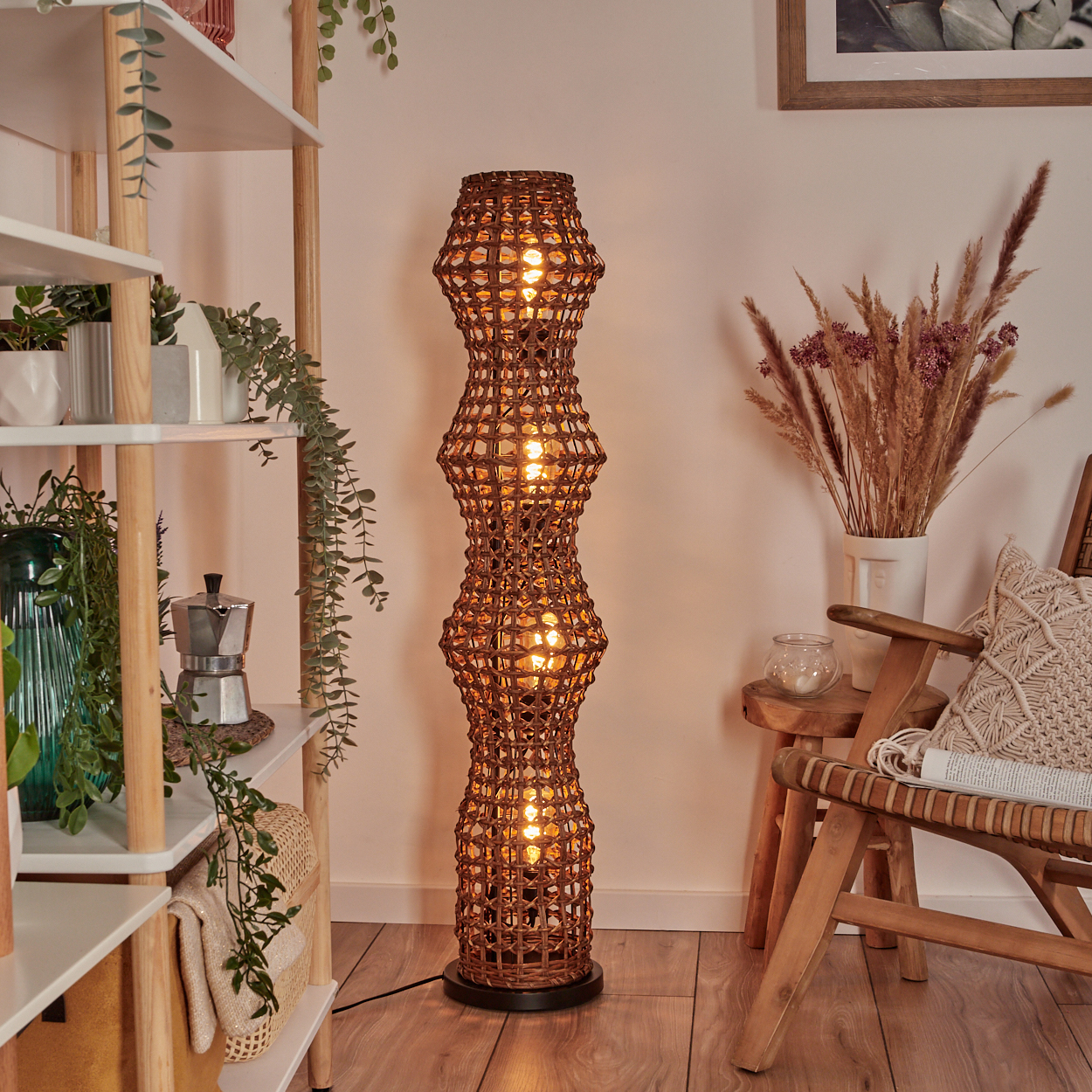 Eine gewebte braune Stehlampe leuchtet, links Pflanzen und Regale, rechts Stuhl mit Kissen und Vase mit Trockenblumen.