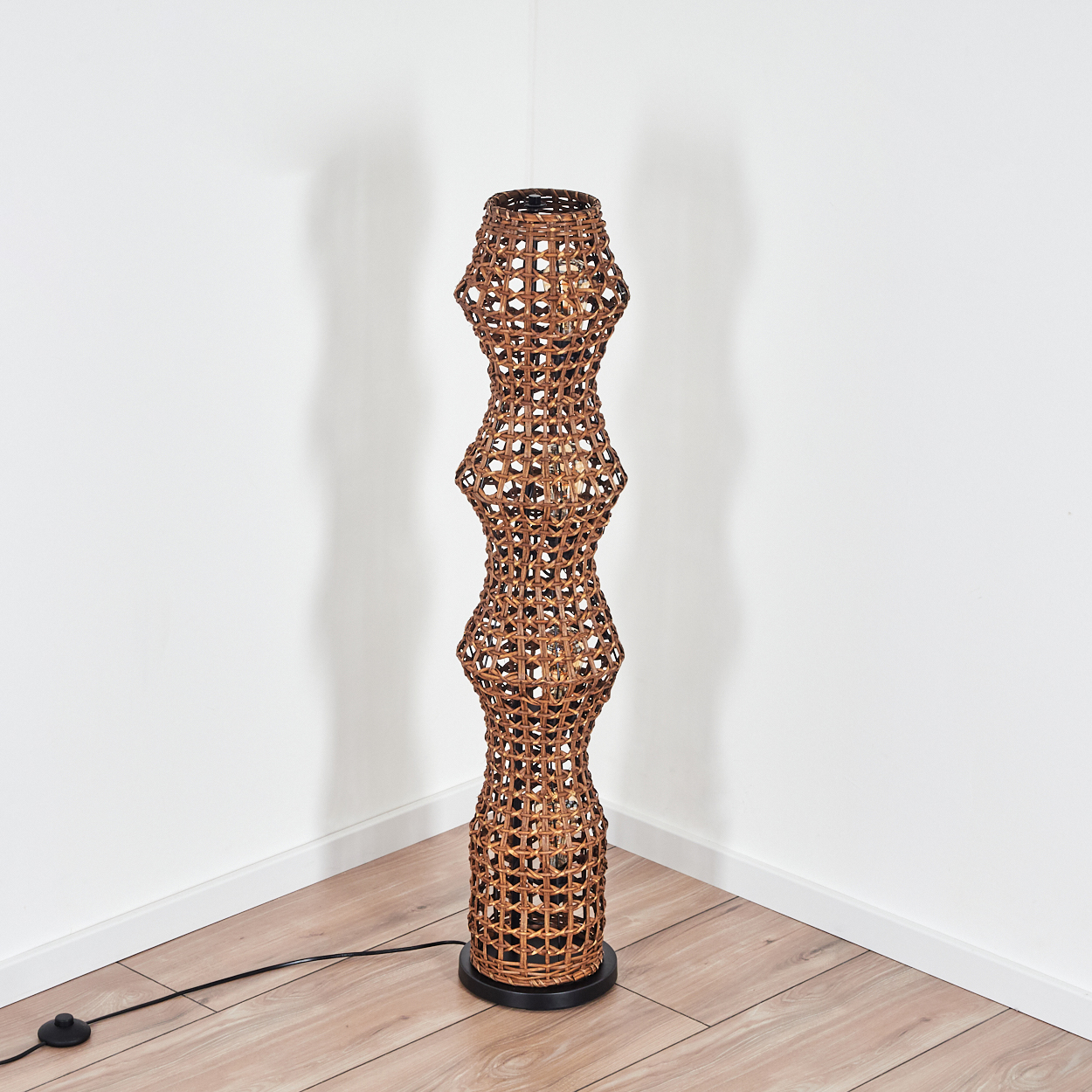 Eine hohe, braune gewebte Lampe steht in einer Ecke auf einem Holzboden. Die Lampe hat einen schwarzen Sockel und ein schwarzes Netzkabel.