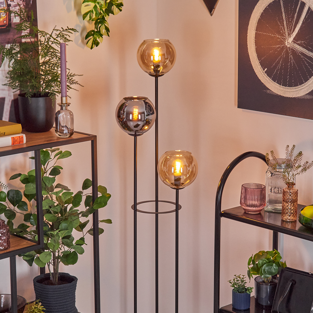 Eine moderne Stehlampe mit drei Glaskugeln. Im Hintergrund Regale mit Pflanzen und Dekorationsartikeln. Ein Fahrradrad-Kunstwerk an der Wand.