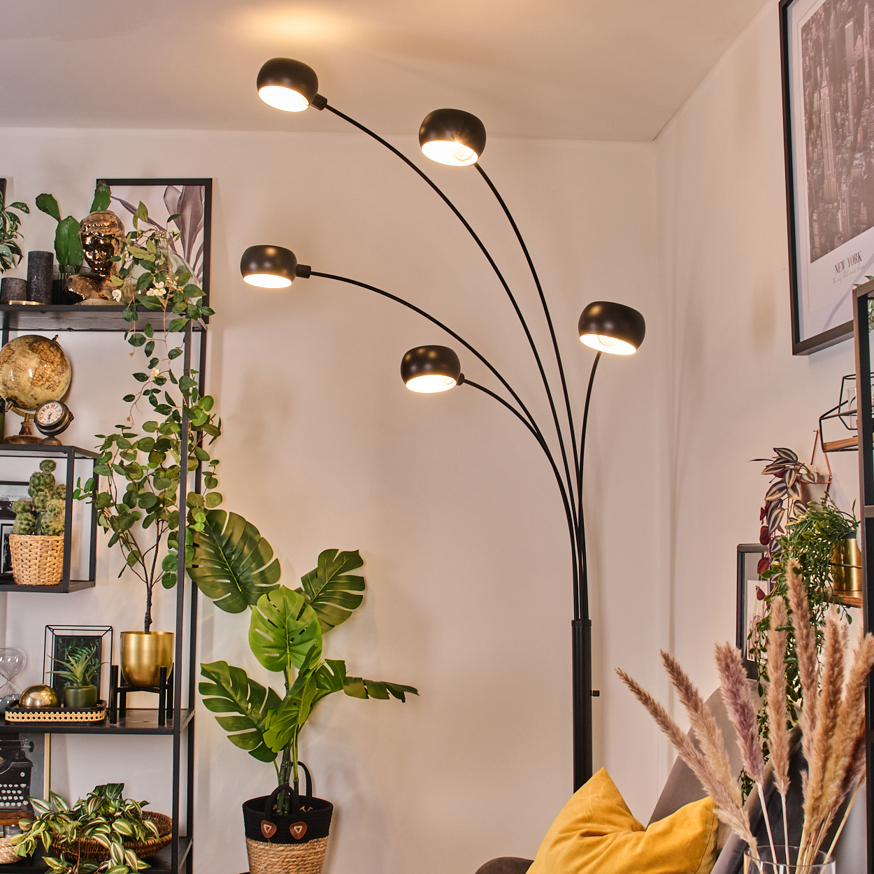 Ein modernes Wohnzimmer mit einer schwarzen Bogenlampe, Pflanzen und einem gelben Kissen. Regale mit Dekoration links.