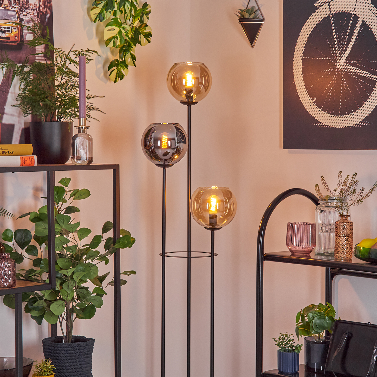 Ein Raum mit einer dreifachen Lampe, Pflanzen und Regalen mit Dekorationen. Ein schwarz-weißes Kunstwerk an der Wand.