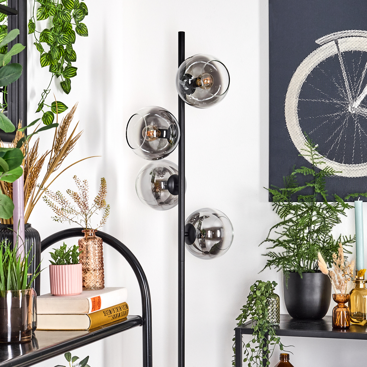 Ein modernes Wohnzimmer mit einer schwarzen Stehlampe mit Glaskugeln. Pflanzen und dekorative Gegenstände stehen auf Regalen und dem Boden.