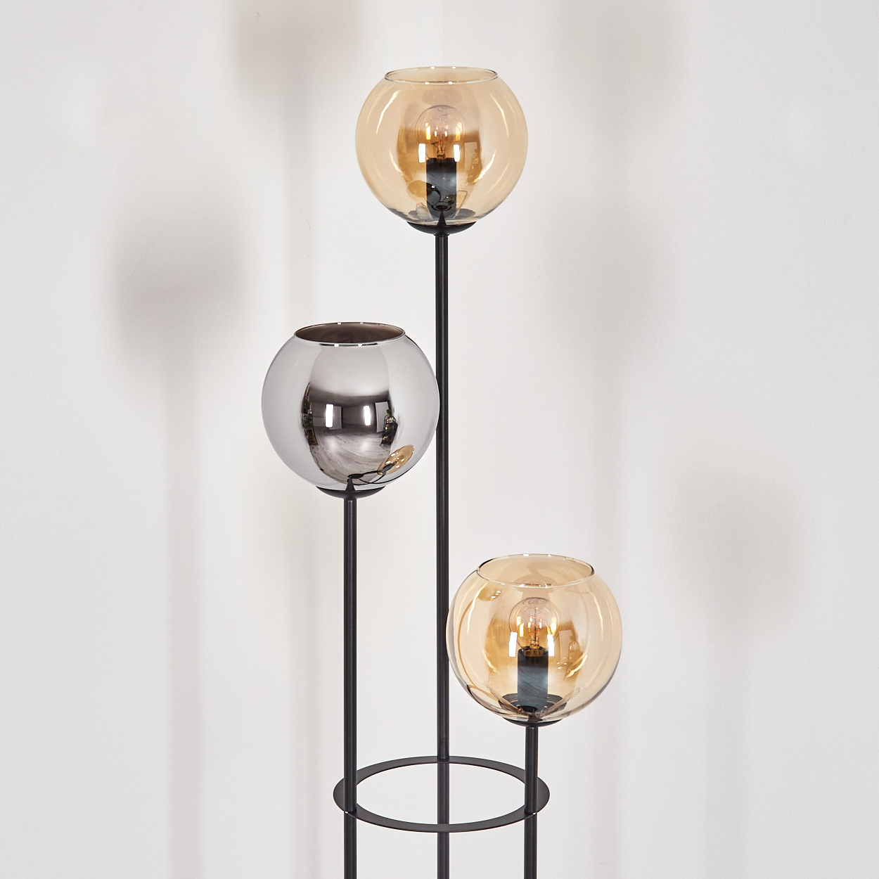 Eine moderne Lampe mit drei Kugelschirmen: zwei bernsteinfarben, einer silberfarben. Schwarzer Metallrahmen. Weißer Wandhintergrund.