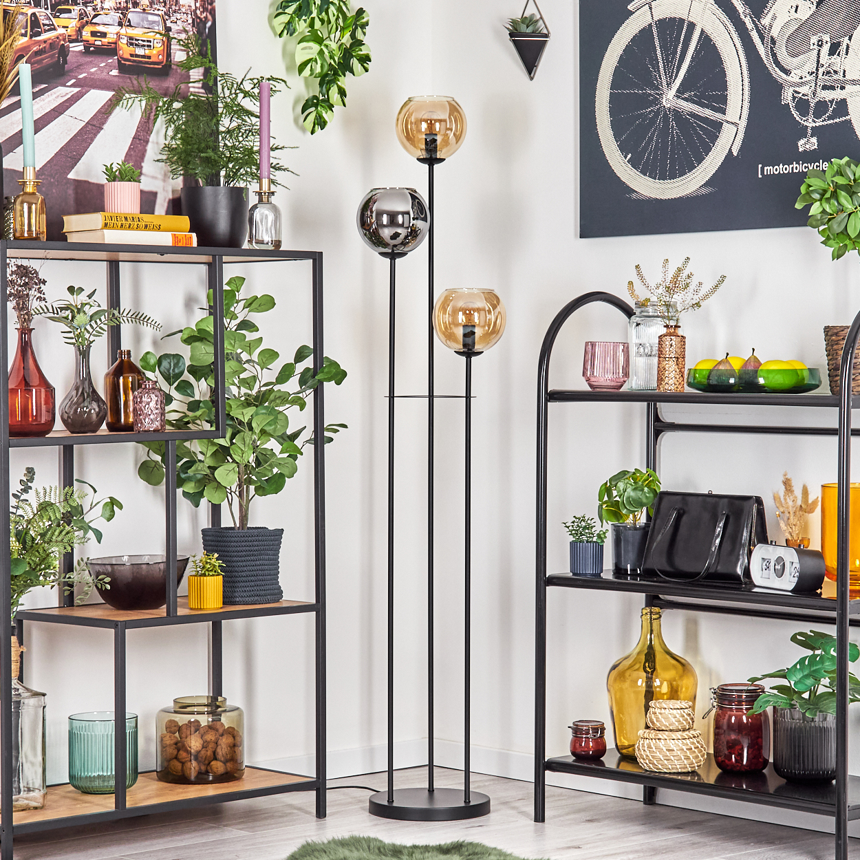 Wohnzimmer mit Regalen, Pflanzen und einer Stehlampe mit drei Glaskugeln. Verschiedene Dekorationsartikel sind ausgestellt.