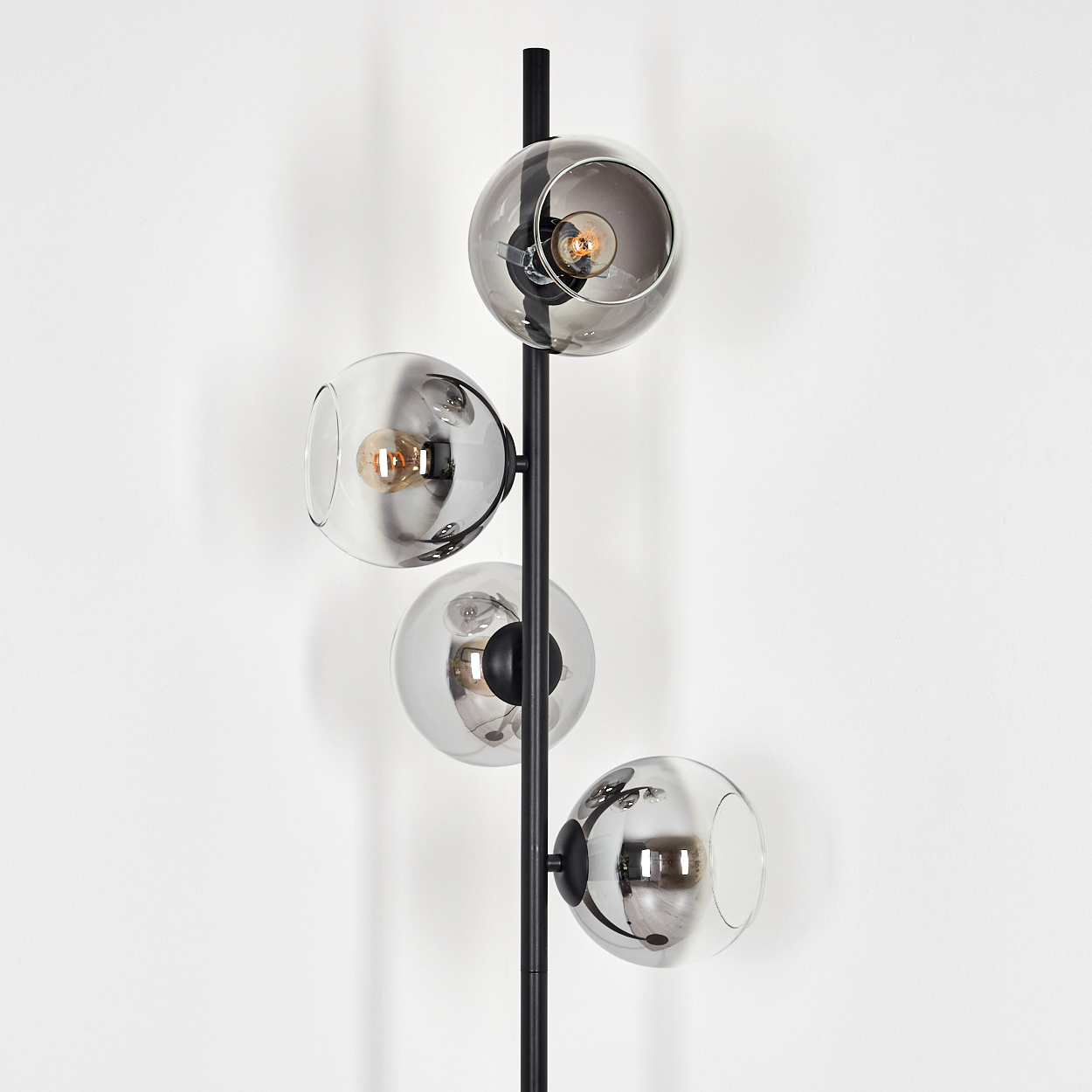 Eine moderne schwarze Stehlampe mit vier kugelförmigen Glasschirmen. Glühbirnen sind in den Schirmen sichtbar. Der Hintergrund ist eine weiße Wand.
