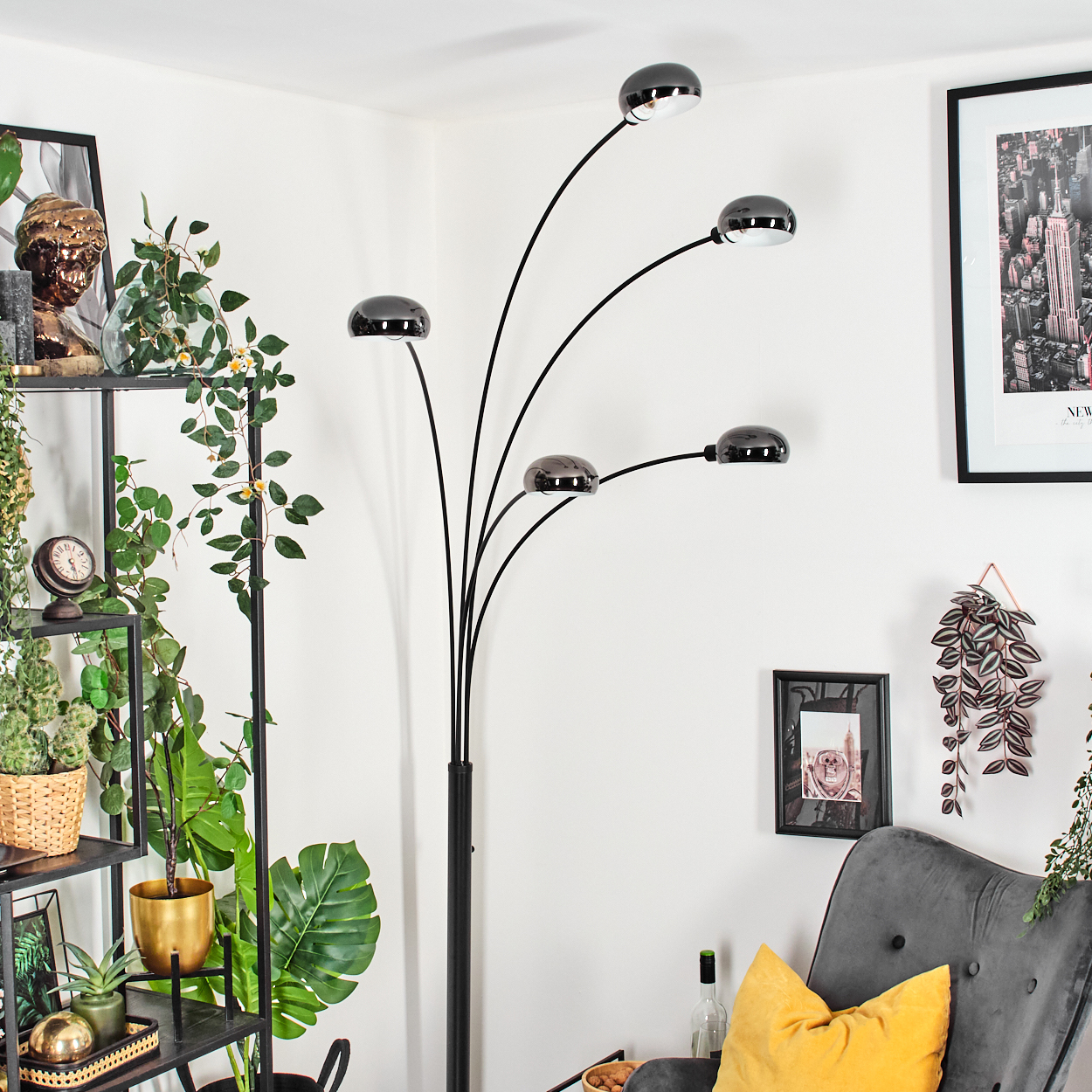 Ein modernes Wohnzimmer mit schwarzer Stehlampe, grauem Sessel und gelbem Kissen. Pflanzen und Bilder schmücken die weißen Wände.
