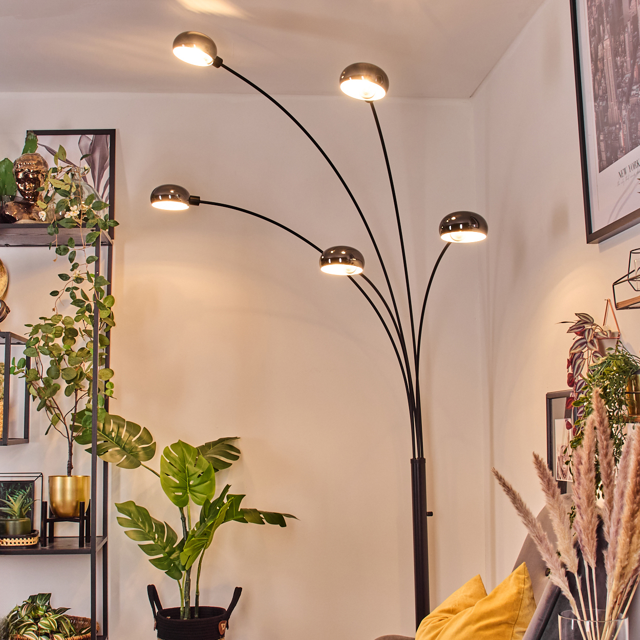 Eine moderne Stehlampe mit fünf Lichtern beleuchtet einen Raum mit Pflanzen, Regalen und einem gelben Kissen auf einem grauen Sofa.