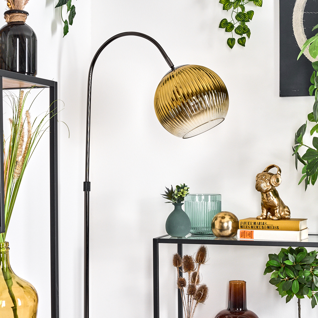 Ein modernes Wohnzimmer mit schwarzem Metallregal, Pflanzen, goldener Lampe und dekorativen Gegenständen wie Vasen und goldener Hundestatue.