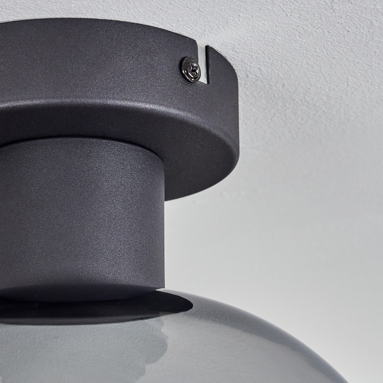 Nahaufnahme einer Deckenlampe. Sie hat einen schwarzen Metallsockel und einen abgerundeten, grauen Glasschirm. Die Decke ist weiß.
