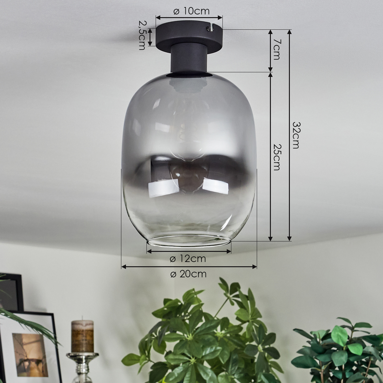 Eine moderne, graue Glasdeckenlampe mit Maßangaben wird gezeigt. Sie hängt über grünen Pflanzen und einer Kerze in einem dekorativen Halter.