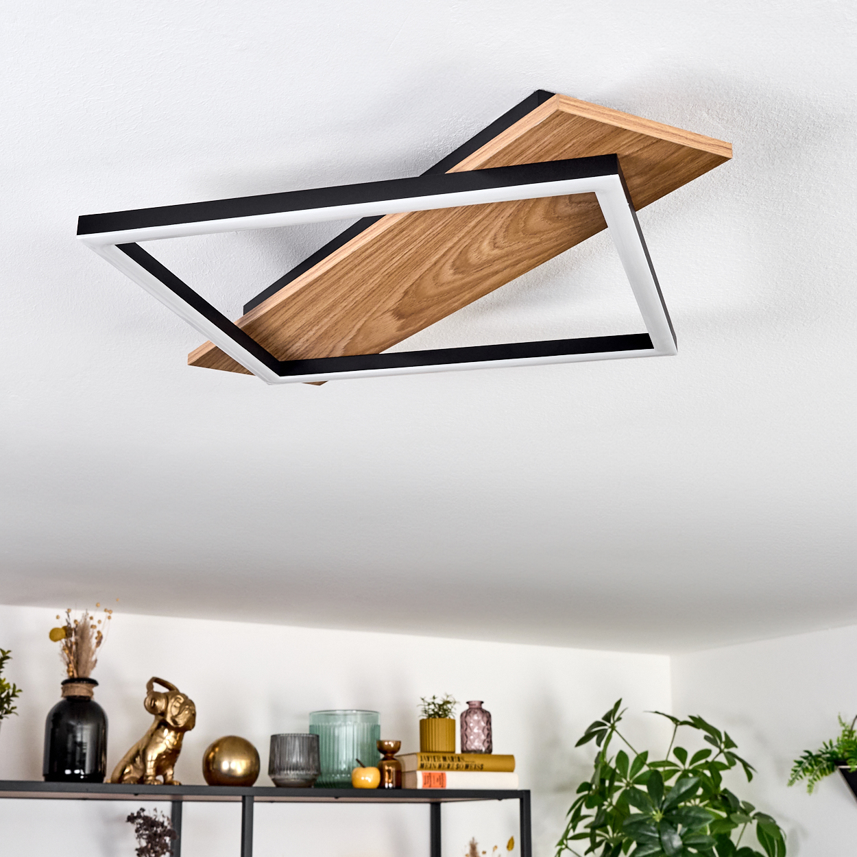 Moderne Deckenleuchte mit Holz und schwarzem Rahmen. Darunter ein Regal mit Dekorationen und Pflanzen. Weiße Decke.