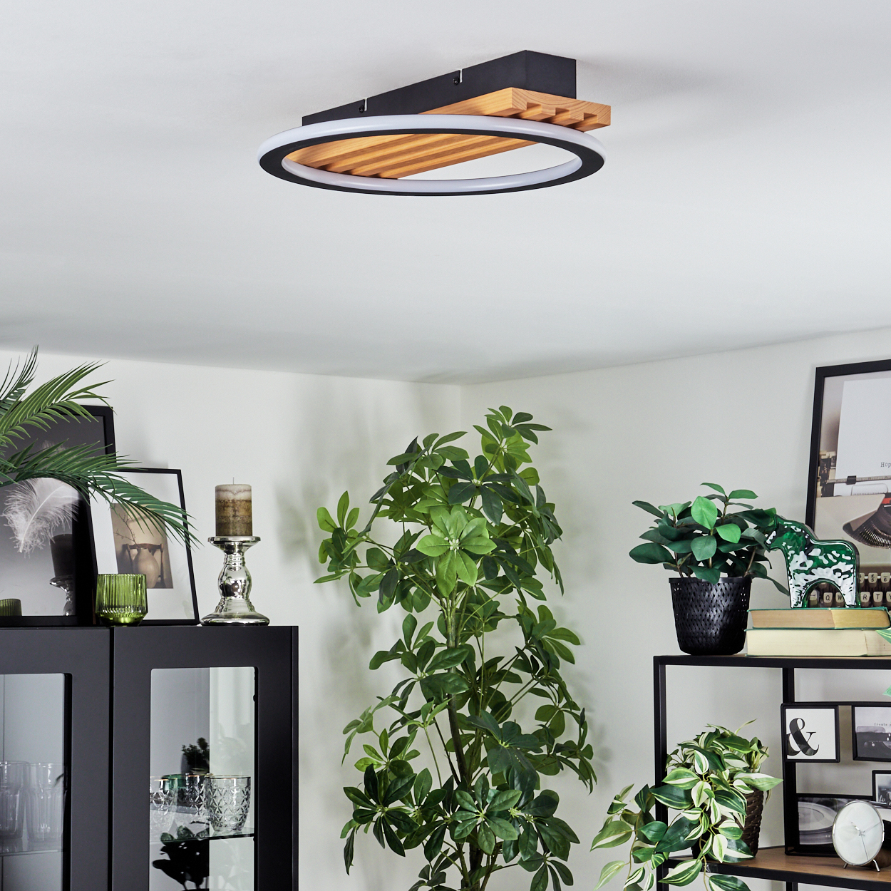 Eine moderne Deckenleuchte mit Holz- und schwarzem Rahmen. Pflanzen und gerahmte Bilder im Vordergrund, weiße Wand im Hintergrund.