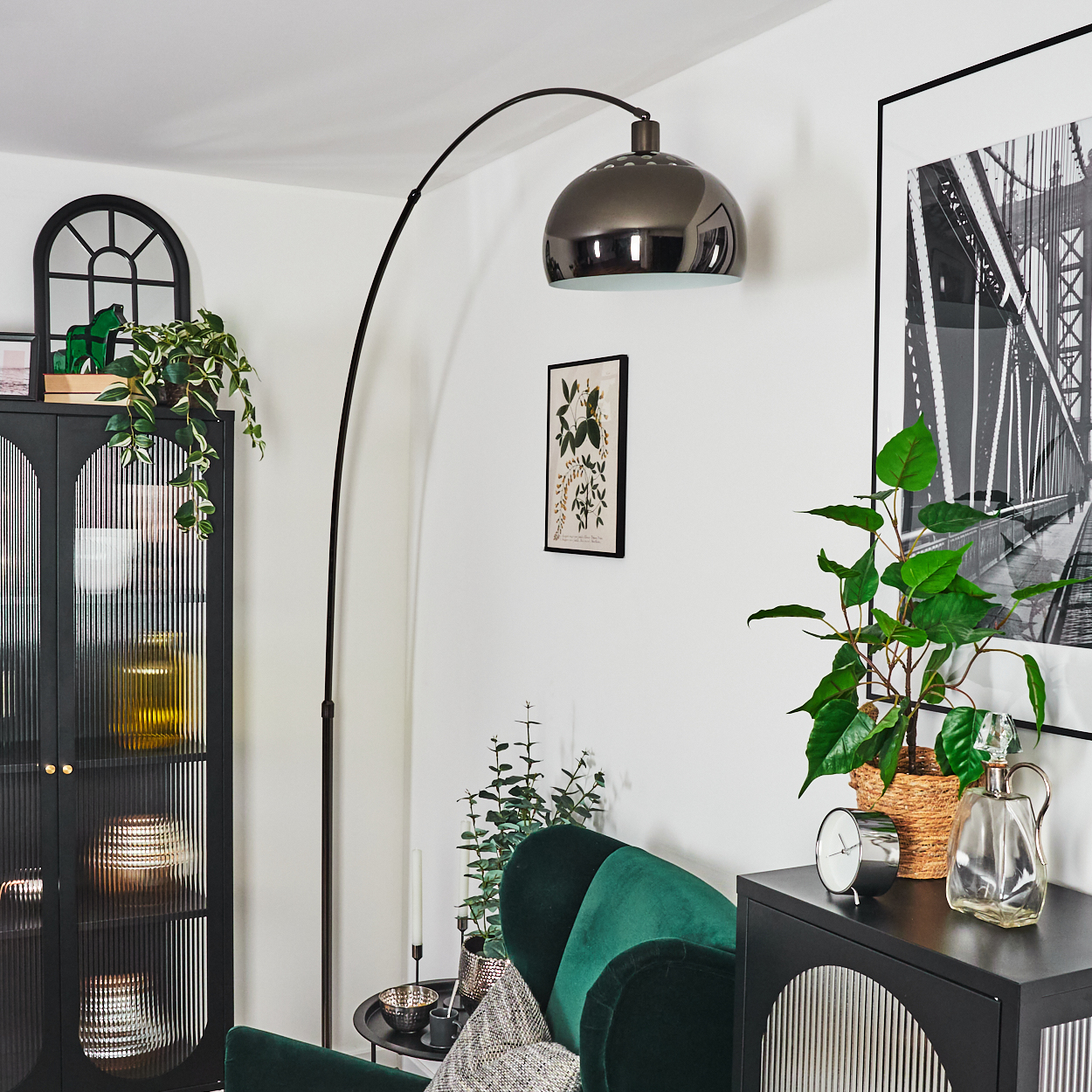 Wohnzimmer mit grünem Samtstuhl, schwarzem Schrank, Bogenlampe und gerahmtem Brückenfoto. Pflanzen und Dekorationsartikel sind vorhanden.